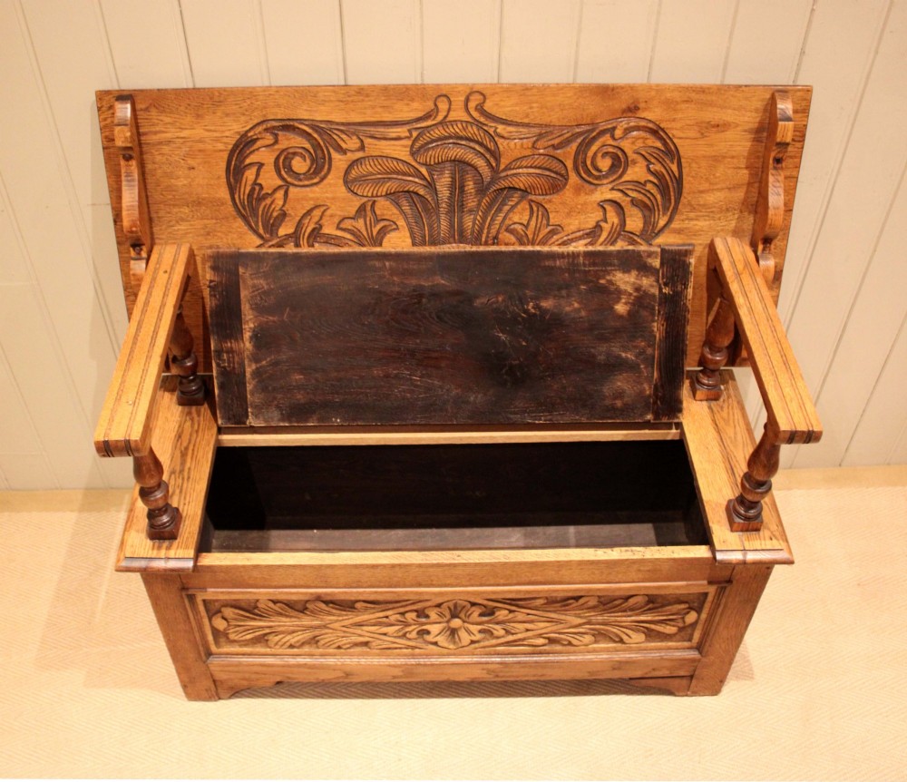 A carved solid oak monks bench featuring a decorative back panel that tilts forward to form a table surface, turned arm supports, and a lift-up storage seat. View V
