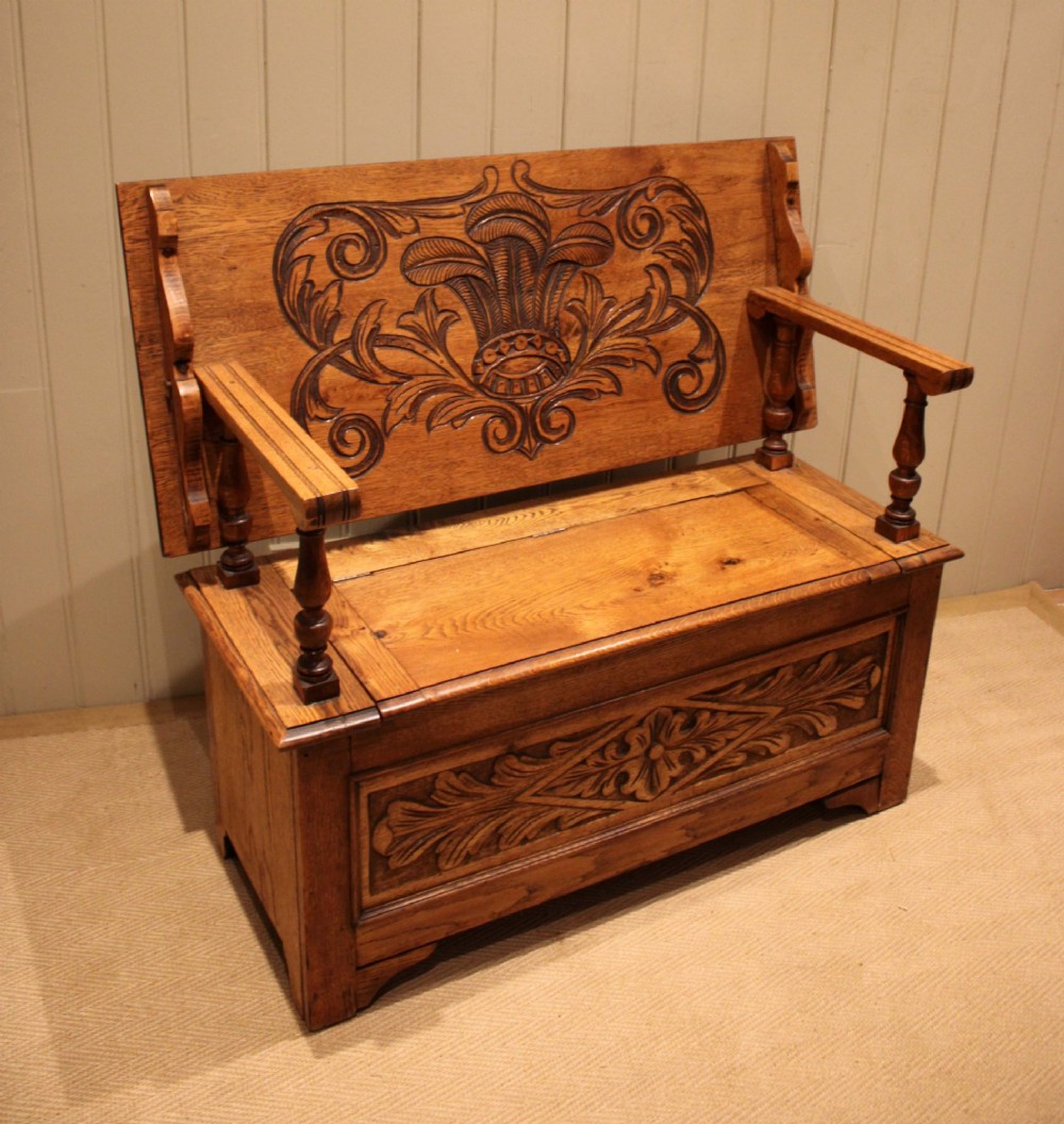 A carved solid oak monks bench featuring a decorative back panel that tilts forward to form a table surface, turned arm supports, and a lift-up storage seat. View VI