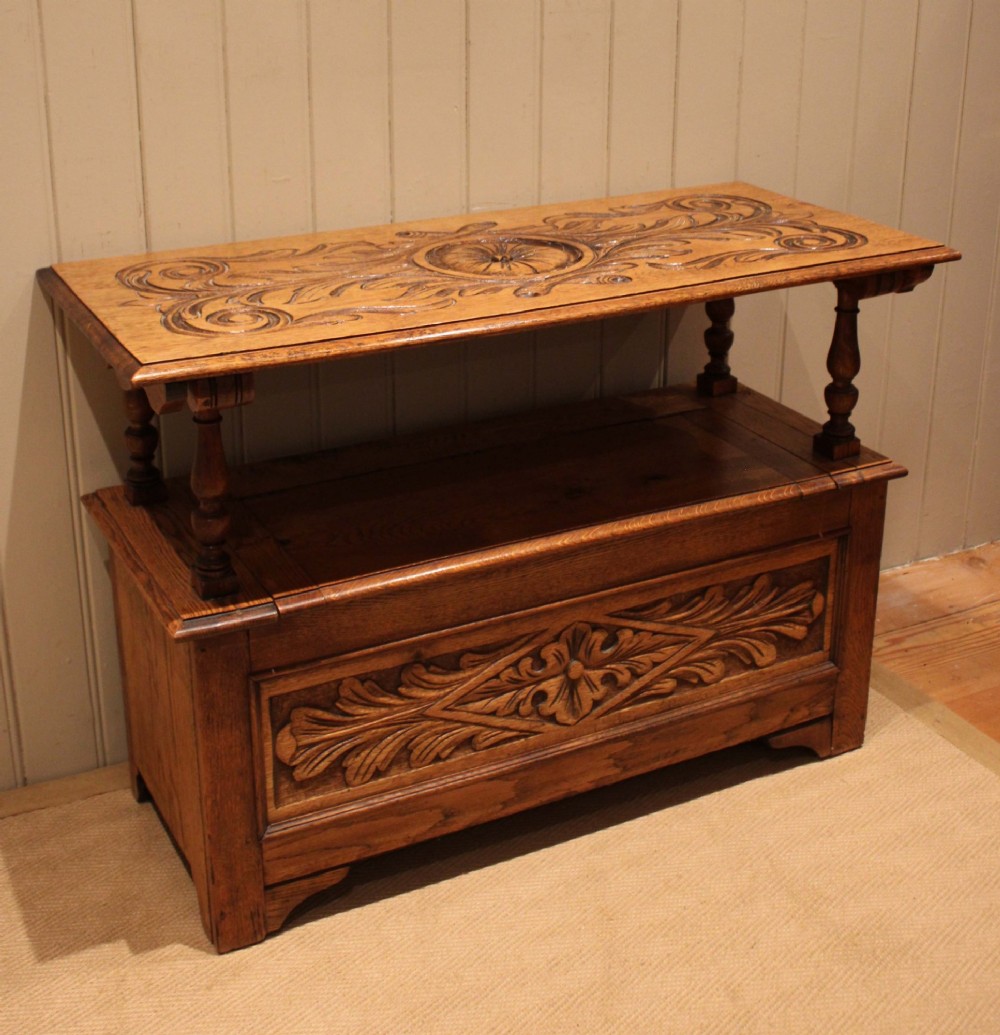 A carved solid oak monks bench featuring a decorative back panel that tilts forward to form a table surface, turned arm supports, and a lift-up storage seat. View VII