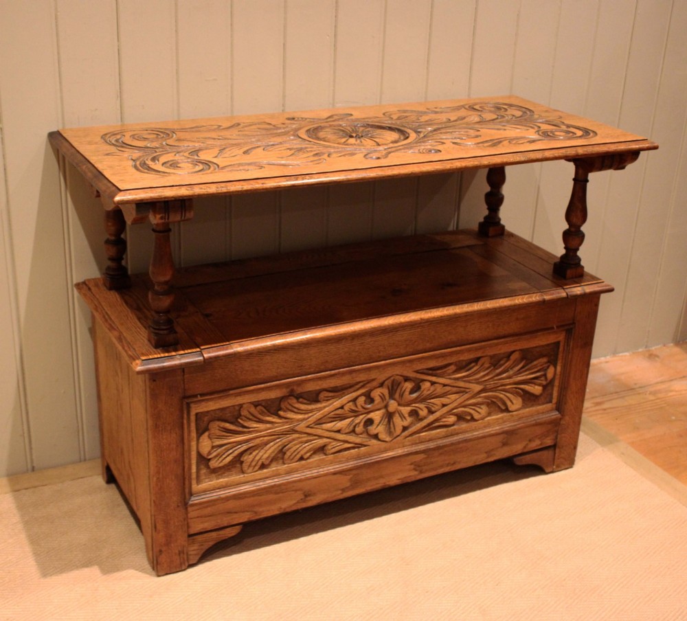 A carved solid oak monks bench featuring a decorative back panel that tilts forward to form a table surface, turned arm supports, and a lift-up storage seat. View IX