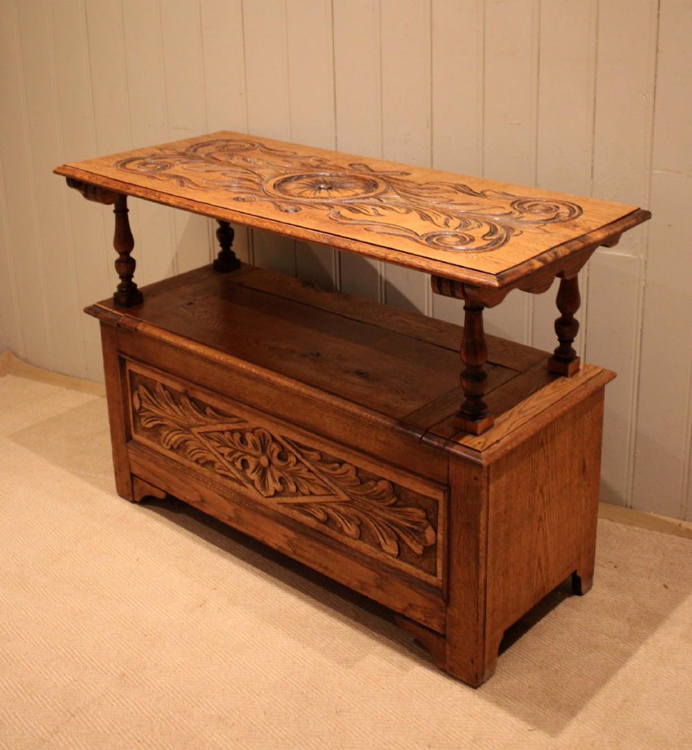 A carved solid oak monks bench featuring a decorative back panel that tilts forward to form a table surface, turned arm supports, and a lift-up storage seat. View X
