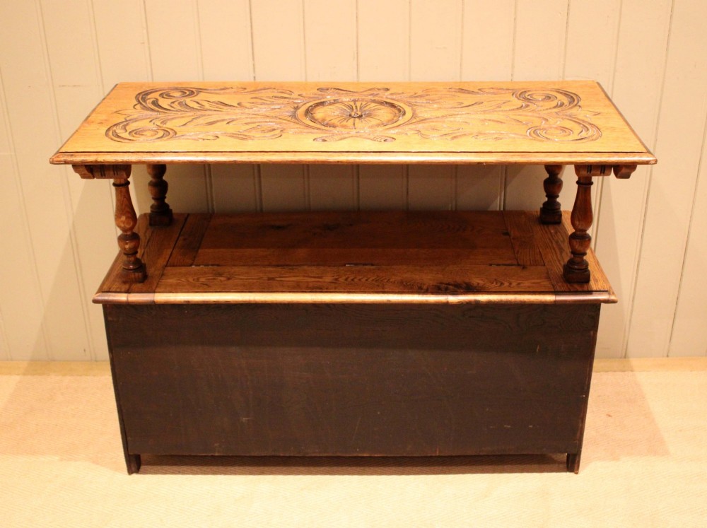A carved solid oak monks bench featuring a decorative back panel that tilts forward to form a table surface, turned arm supports, and a lift-up storage seat. View XII