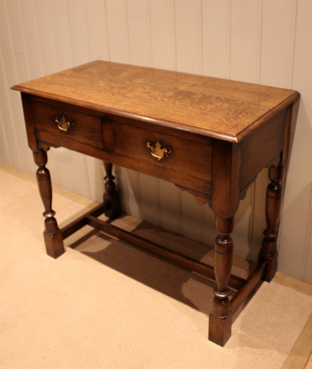 A rectangular solid oak side table featuring a quarter-sawn top, two frieze drawers with brass handles, turned legs, and an H-stretcher base. View X