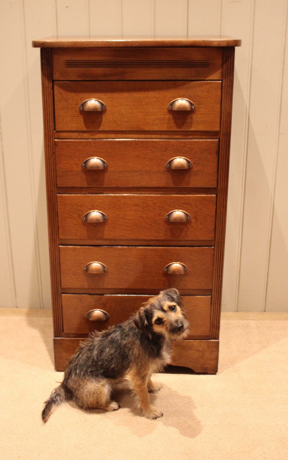 A tall oak chest of drawers featuring five drawers with metal cup handles and reeded side detailing. View X