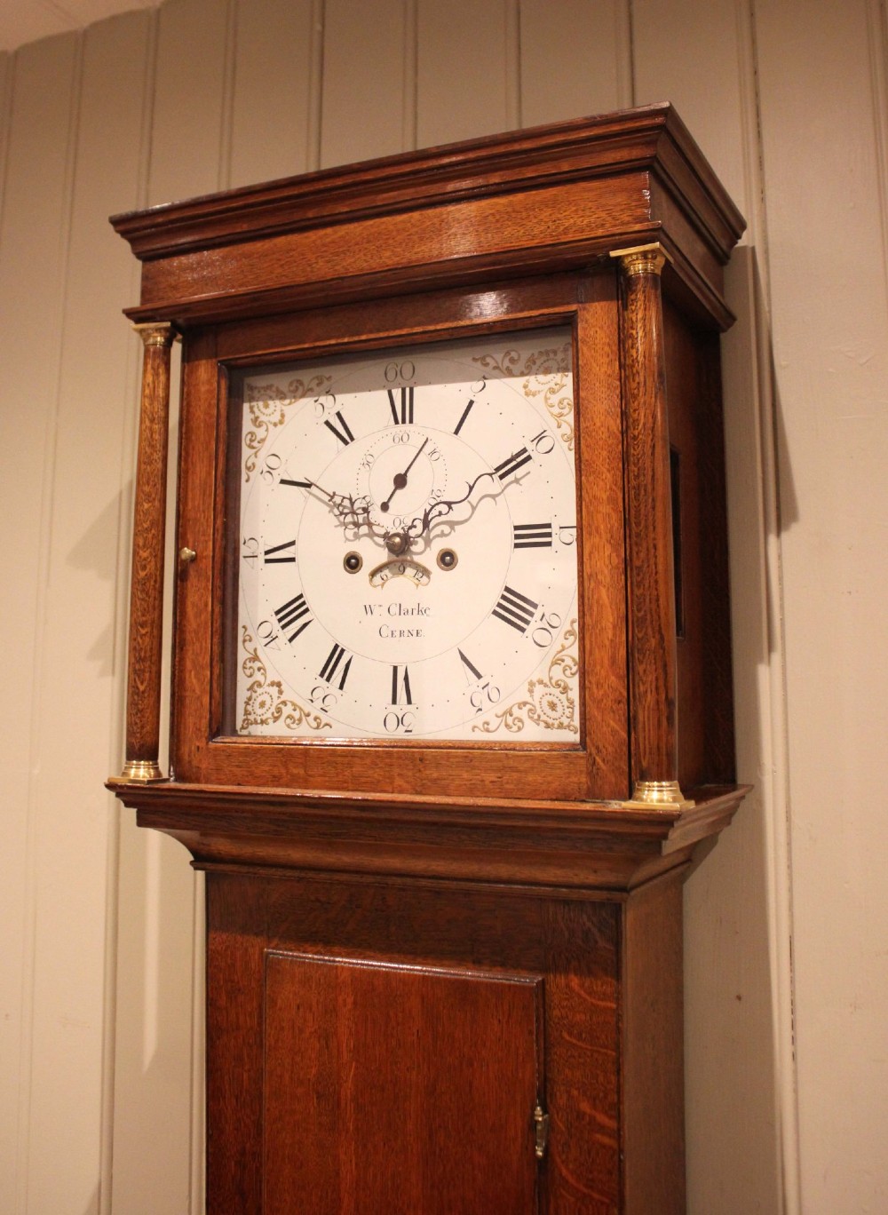 A tall late Georgian oak longcase clock featuring a square painted dial, turned hood columns, and a polished wood trunk. View II