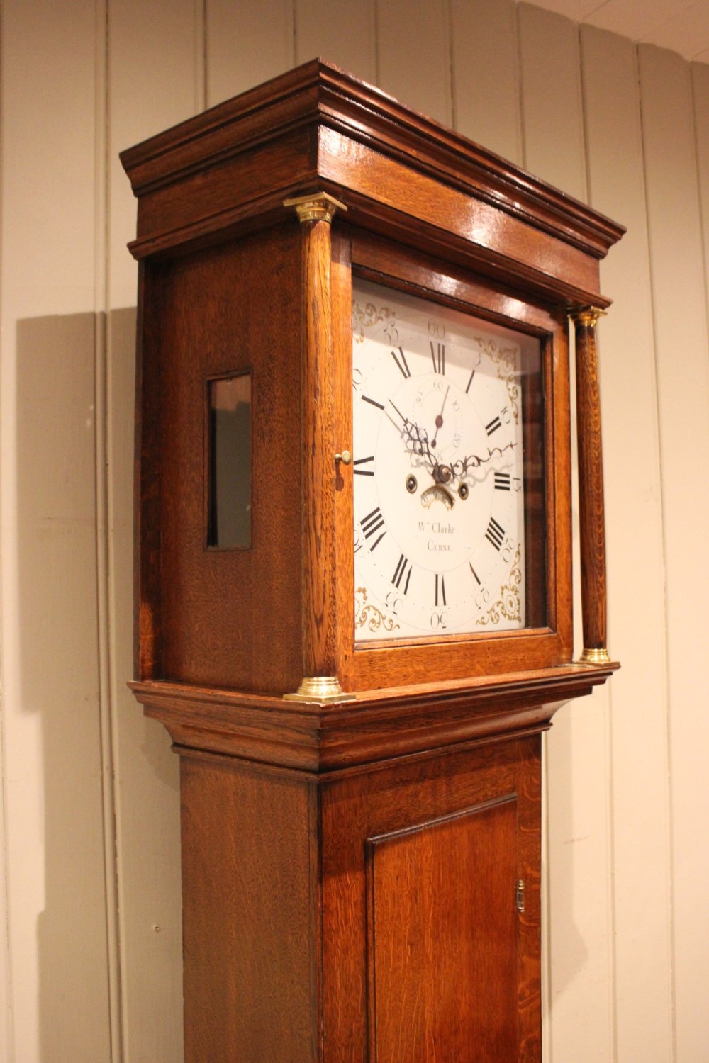 A tall late Georgian oak longcase clock featuring a square painted dial, turned hood columns, and a polished wood trunk. View VI