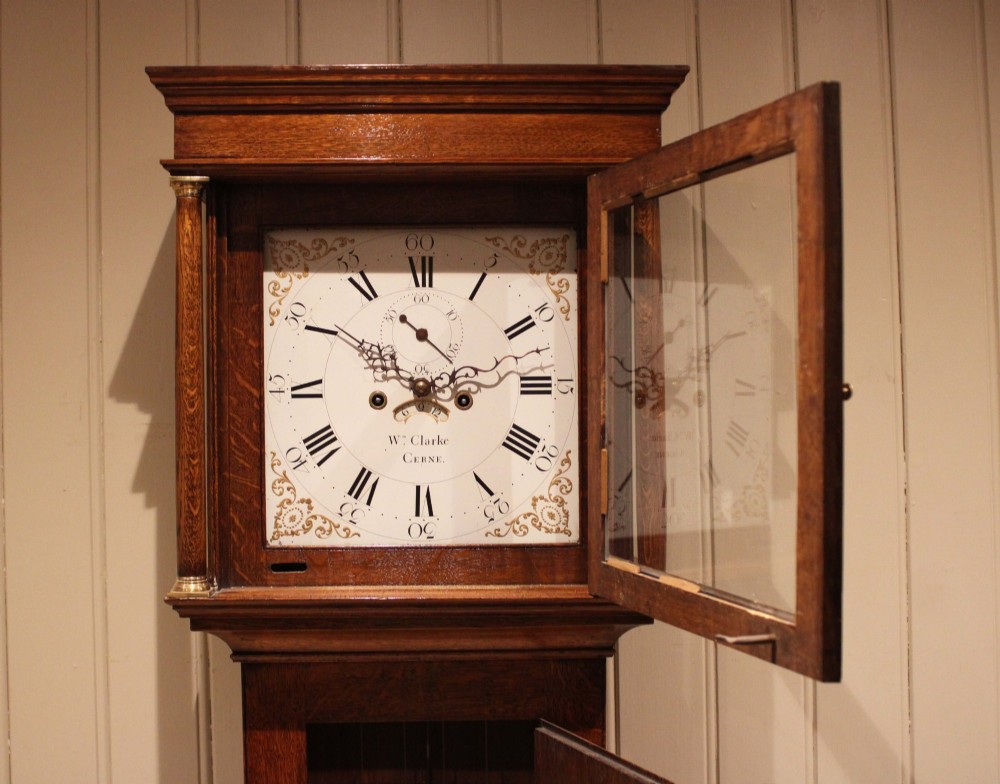 A tall late Georgian oak longcase clock featuring a square painted dial, turned hood columns, and a polished wood trunk. View VII