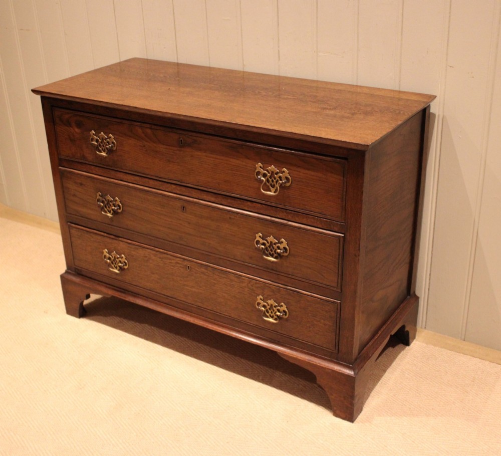 A low Georgian style dark oak chest featuring three full-width drawers with ornate brass handles, set on bracket feet. View III