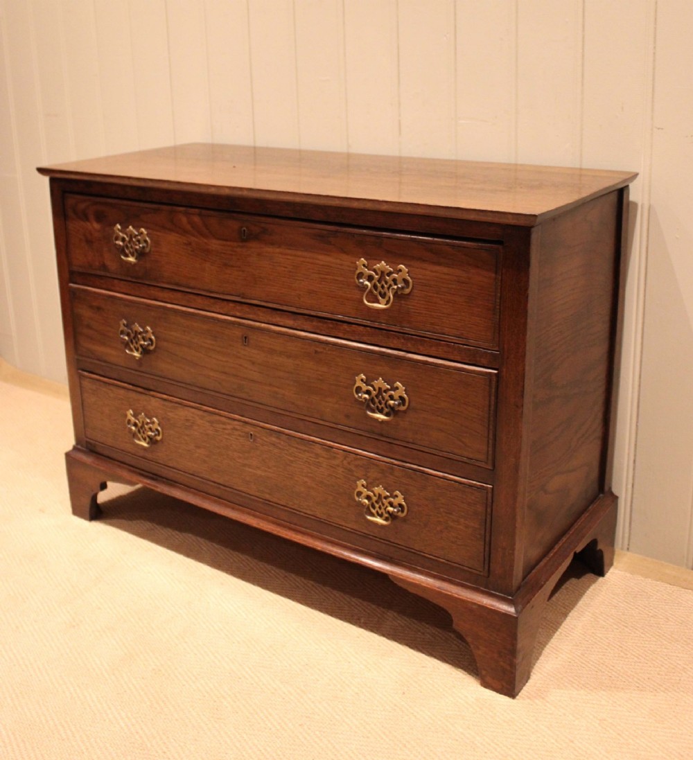 A low Georgian style dark oak chest featuring three full-width drawers with ornate brass handles, set on bracket feet. View V