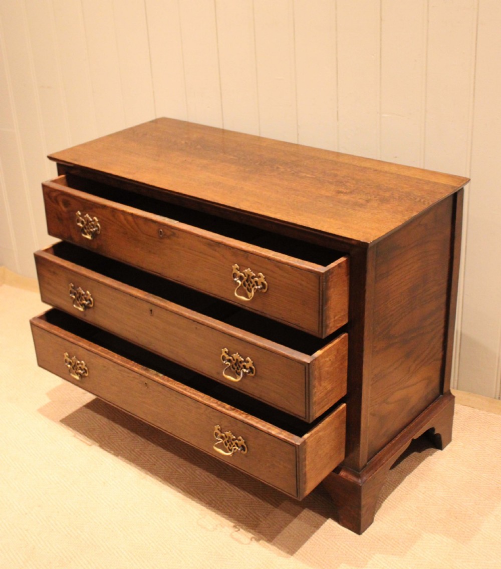 A low Georgian style dark oak chest featuring three full-width drawers with ornate brass handles, set on bracket feet. View XI