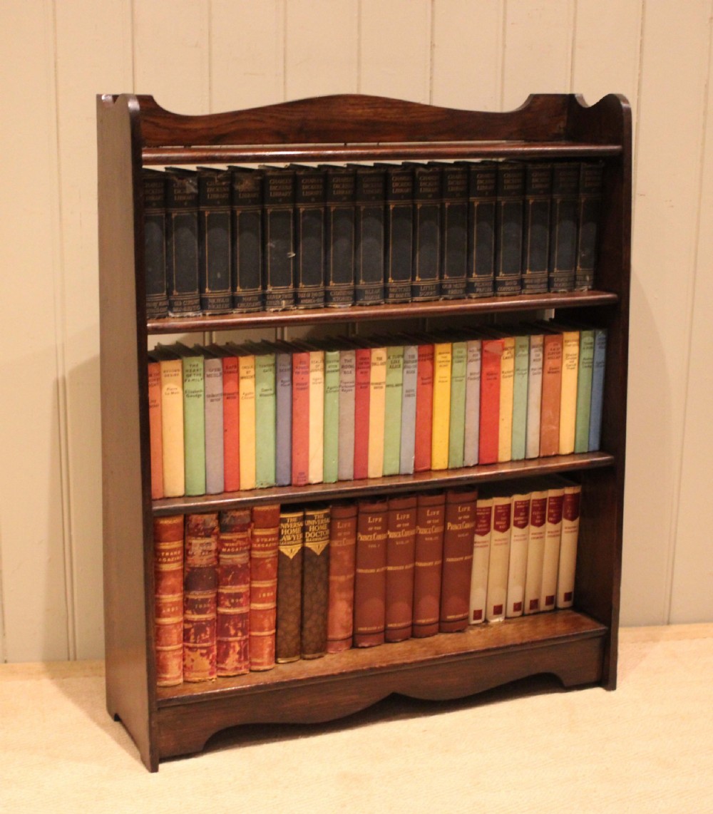 A dark oak open bookcase with three shelves filled with books, featuring a gently curved top cresting and shaped apron.