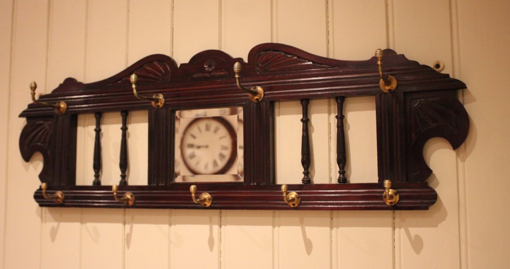 A late 19th-century dark mahogany wall-mounted coat rack featuring turned wooden spindles, a central square mirror, carved crest details, and ten brass hooks.