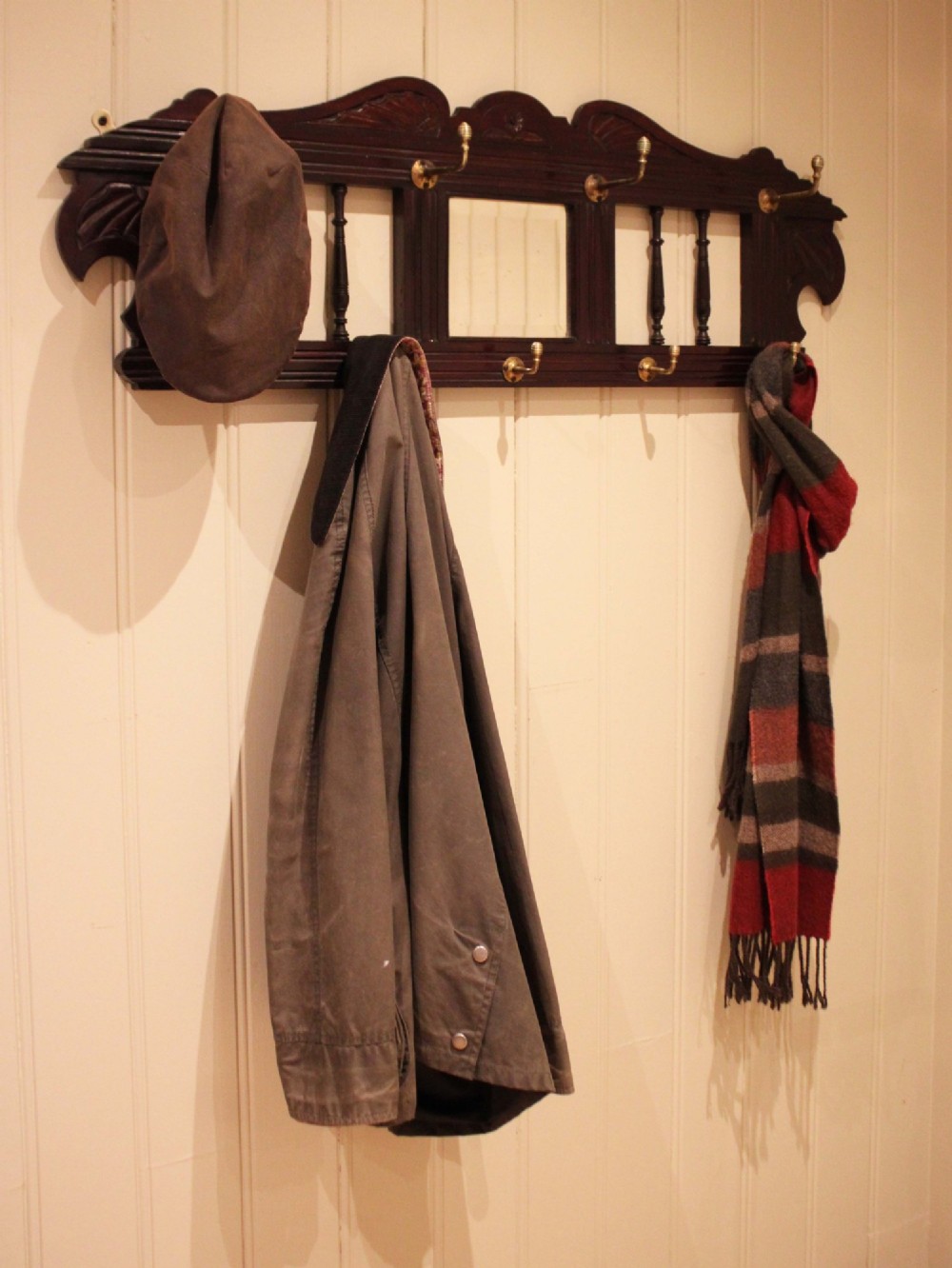A late 19th-century dark mahogany wall-mounted coat rack featuring turned wooden spindles, a central square mirror, carved crest details, and ten brass hooks. View II
