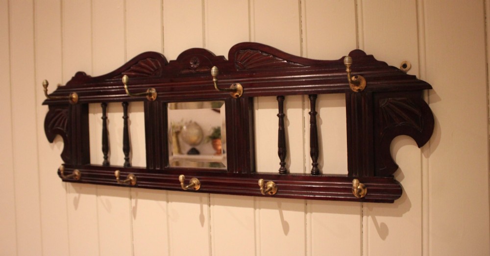 A late 19th-century dark mahogany wall-mounted coat rack featuring turned wooden spindles, a central square mirror, carved crest details, and ten brass hooks. View V