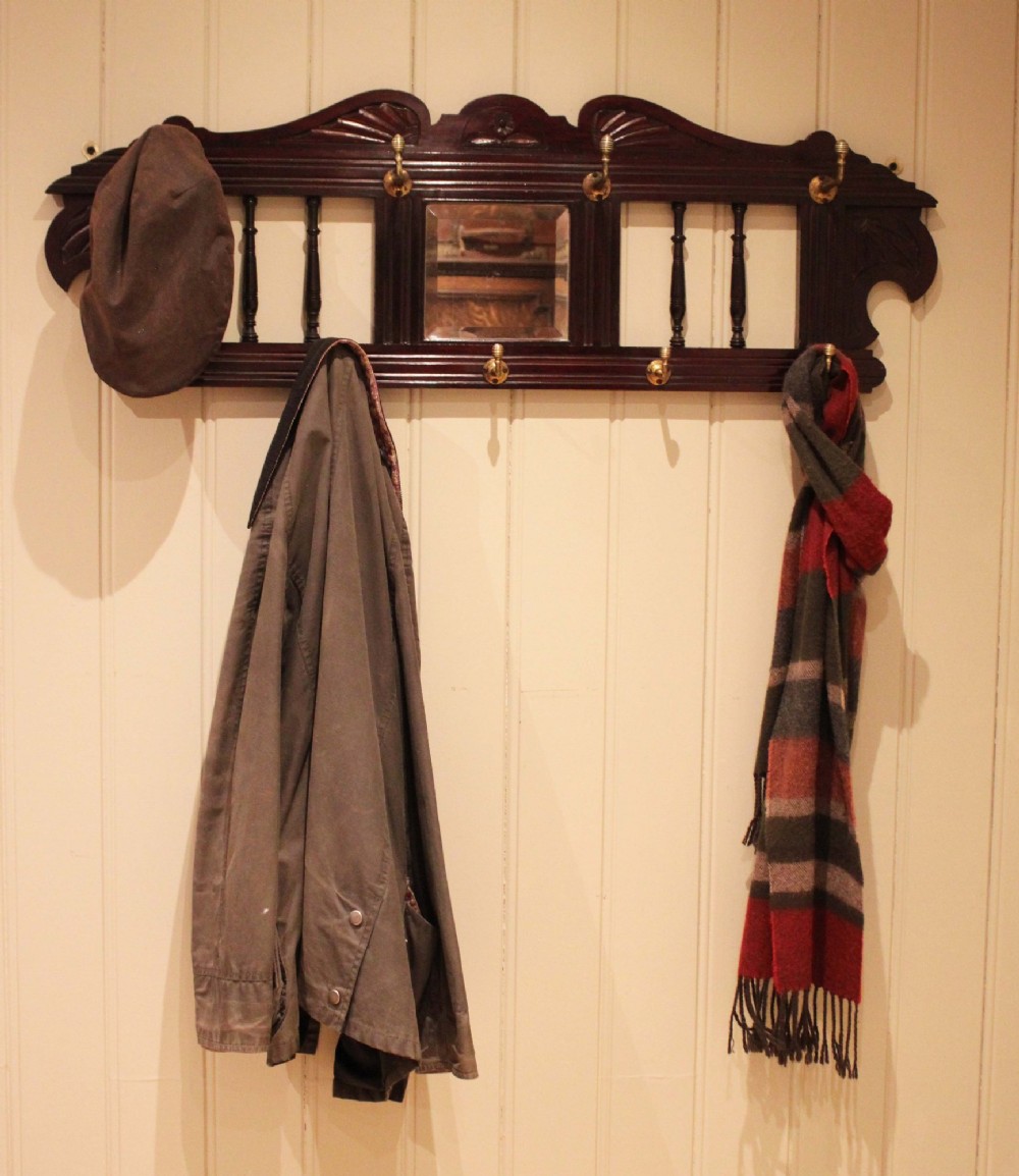 A late 19th-century dark mahogany wall-mounted coat rack featuring turned wooden spindles, a central square mirror, carved crest details, and ten brass hooks. View VI