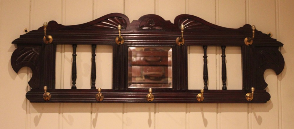 A late 19th-century dark mahogany wall-mounted coat rack featuring turned wooden spindles, a central square mirror, carved crest details, and ten brass hooks. View X