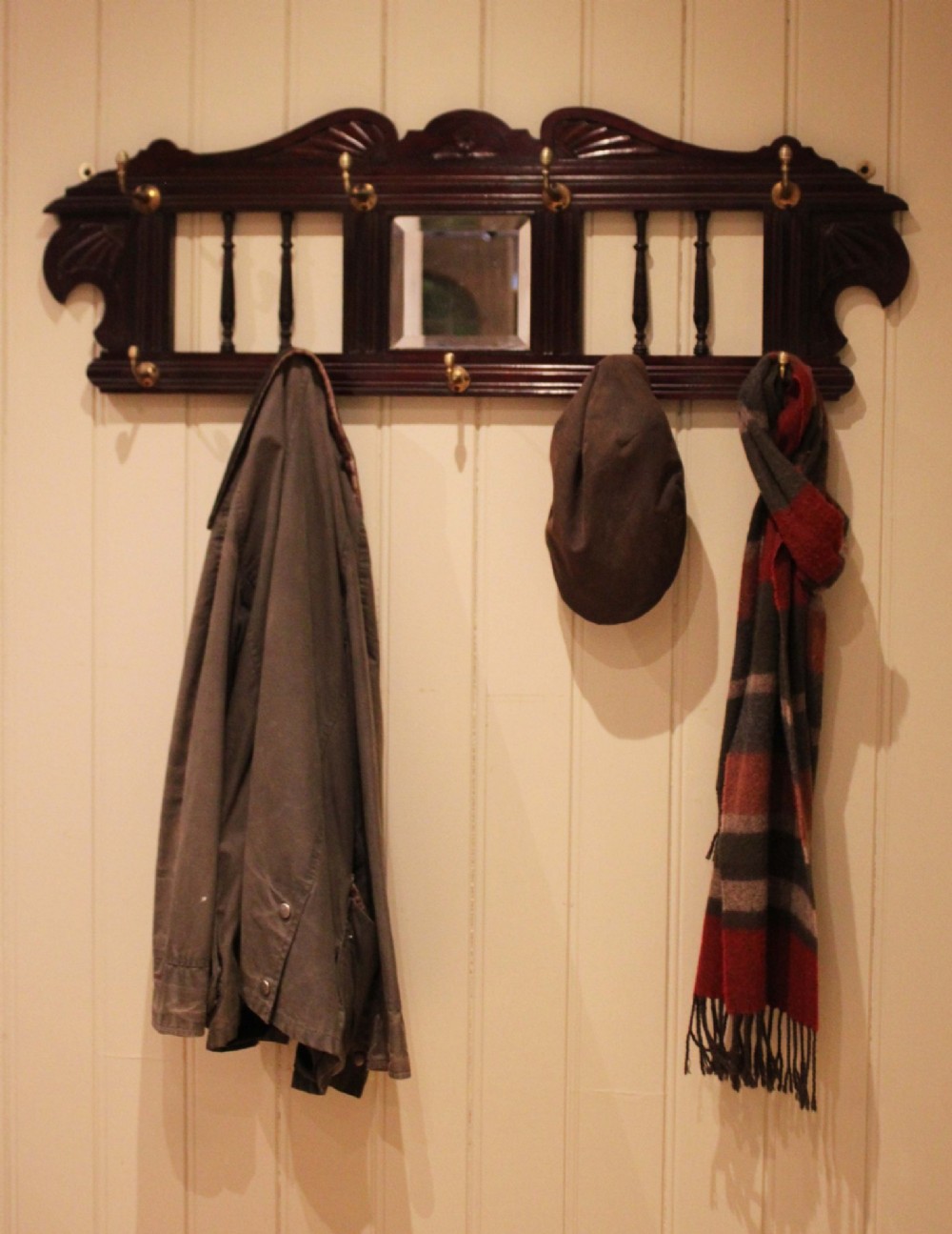 A late 19th-century dark mahogany wall-mounted coat rack featuring turned wooden spindles, a central square mirror, carved crest details, and ten brass hooks. View XII