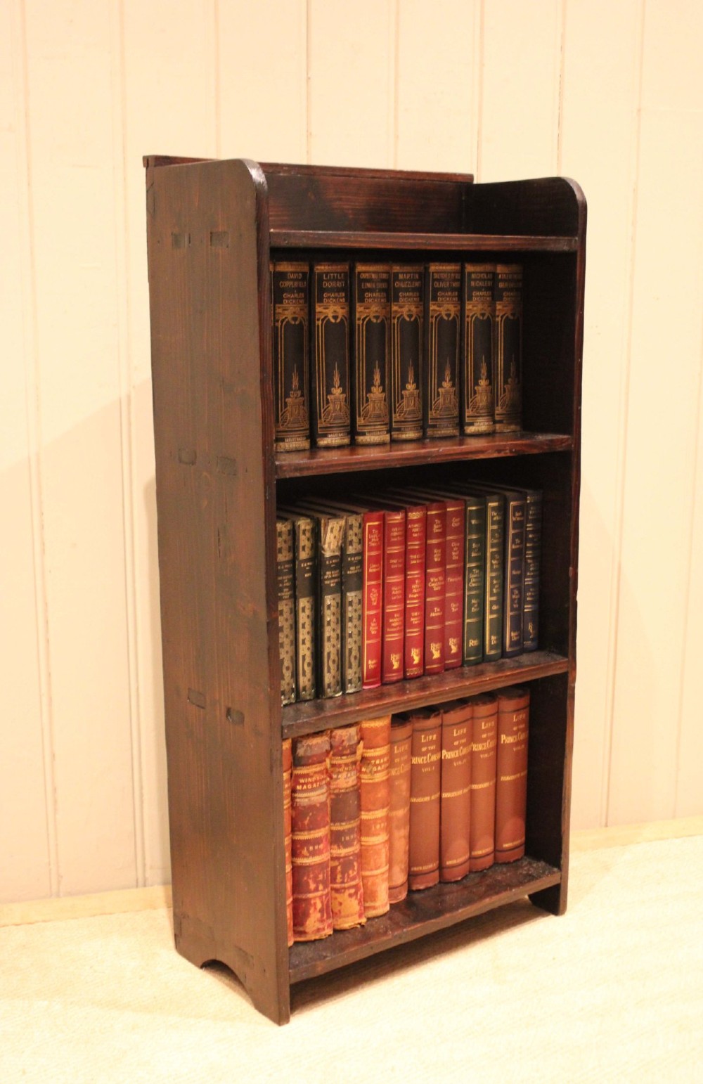 A small dark pine open bookcase featuring three fixed shelves, solid side panels with visible tenon joinery, and a simple shaped gallery top.