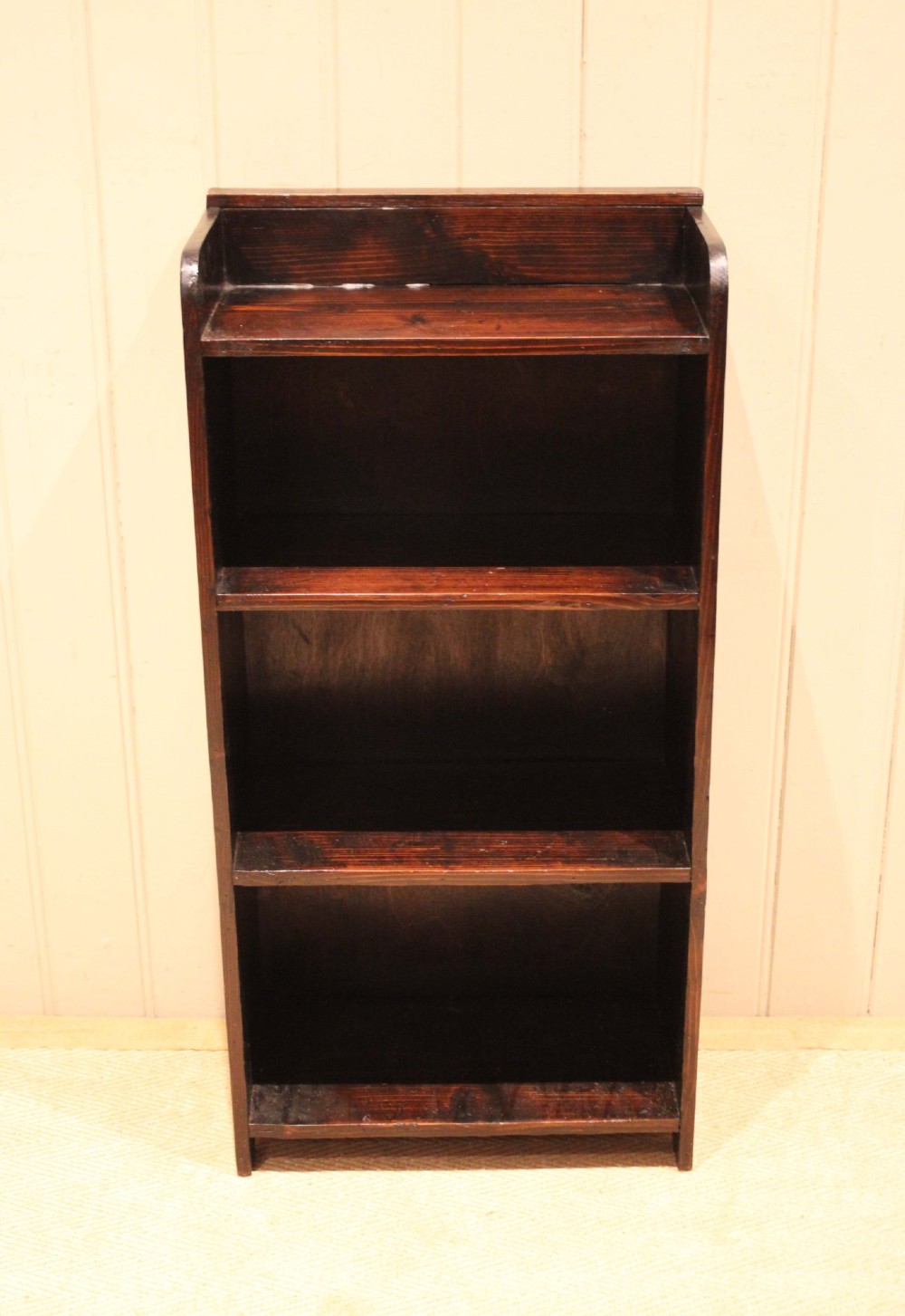 A small dark pine open bookcase featuring three fixed shelves, solid side panels with visible tenon joinery, and a simple shaped gallery top. View II