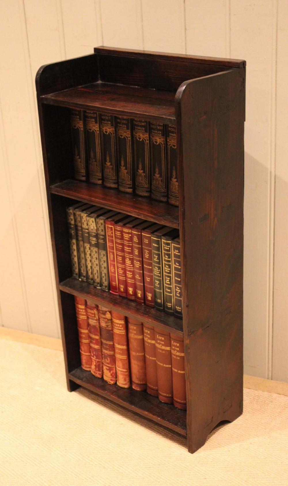 A small dark pine open bookcase featuring three fixed shelves, solid side panels with visible tenon joinery, and a simple shaped gallery top. View III