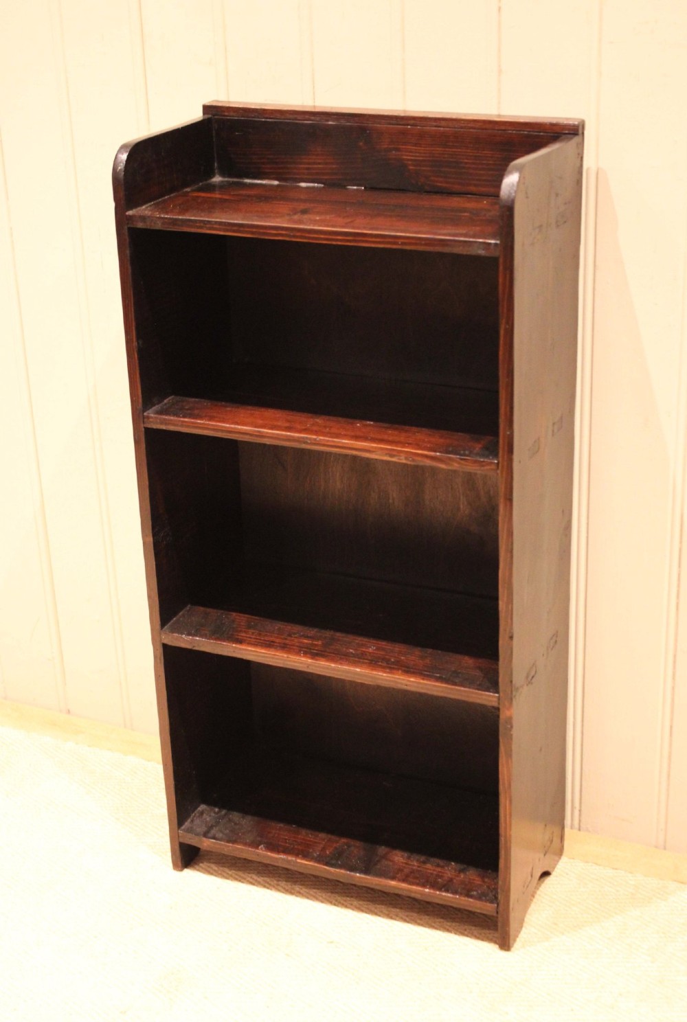 A small dark pine open bookcase featuring three fixed shelves, solid side panels with visible tenon joinery, and a simple shaped gallery top. View IV