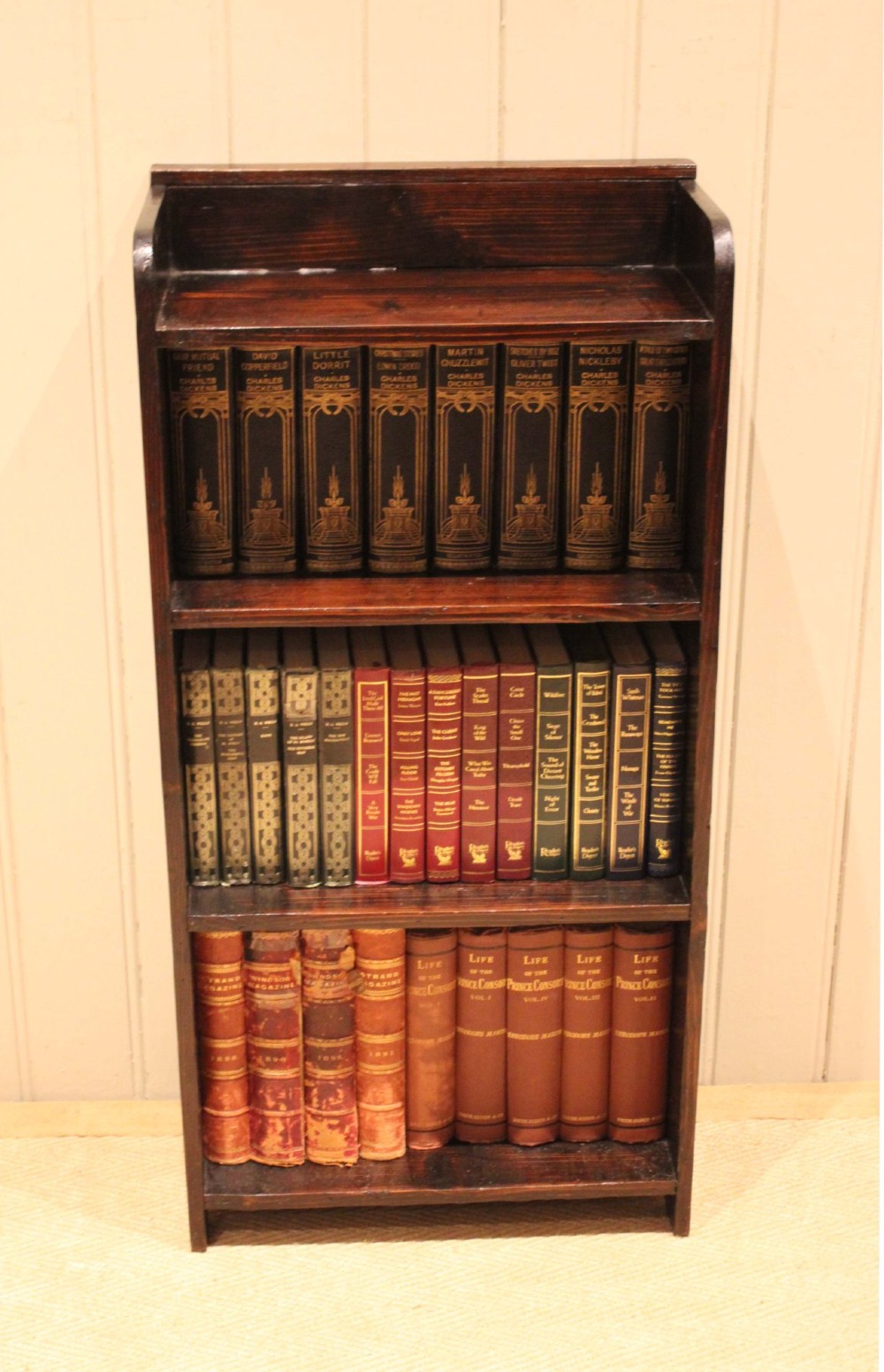 A small dark pine open bookcase featuring three fixed shelves, solid side panels with visible tenon joinery, and a simple shaped gallery top. View V