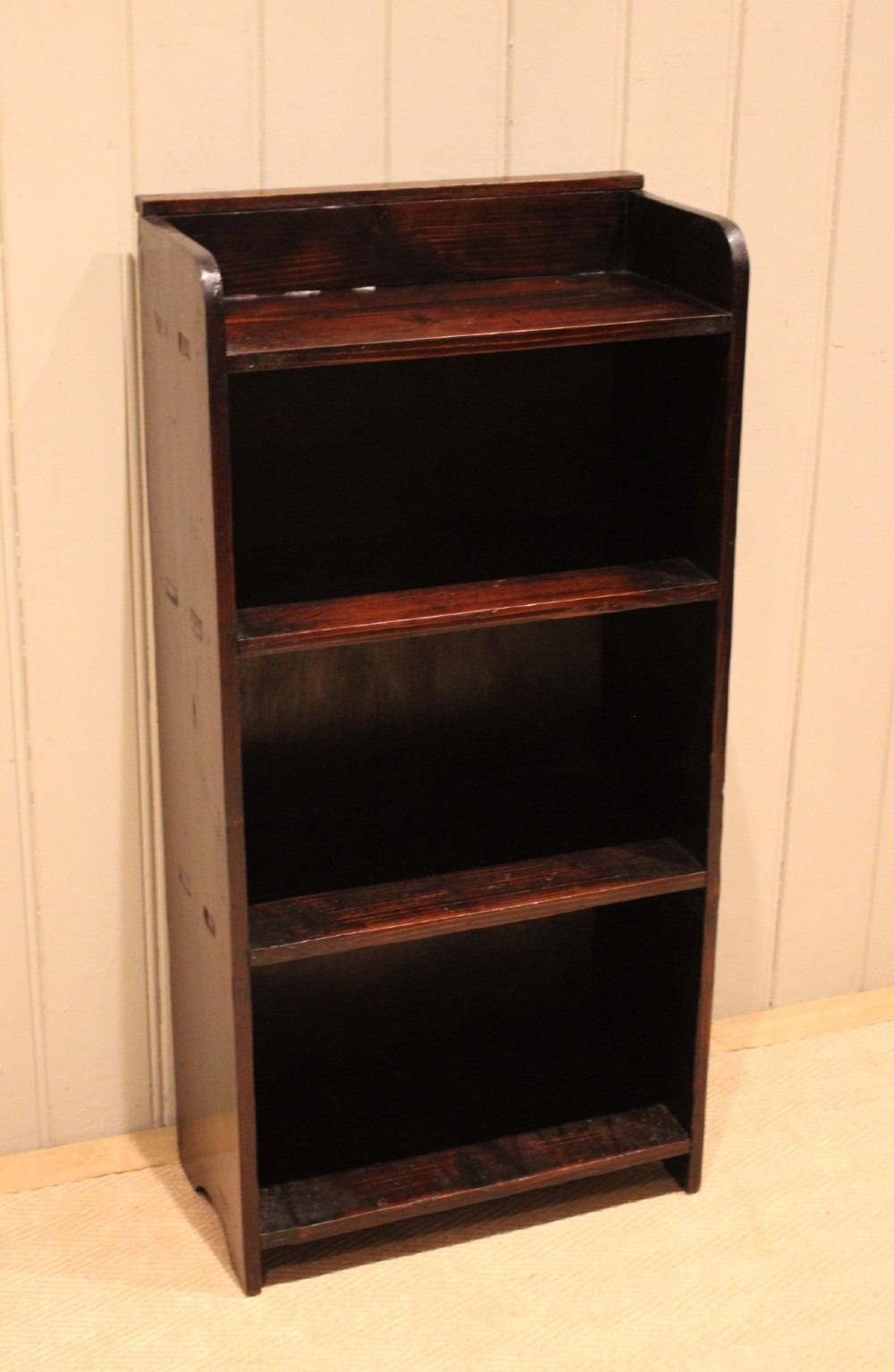 A small dark pine open bookcase featuring three fixed shelves, solid side panels with visible tenon joinery, and a simple shaped gallery top. View VI
