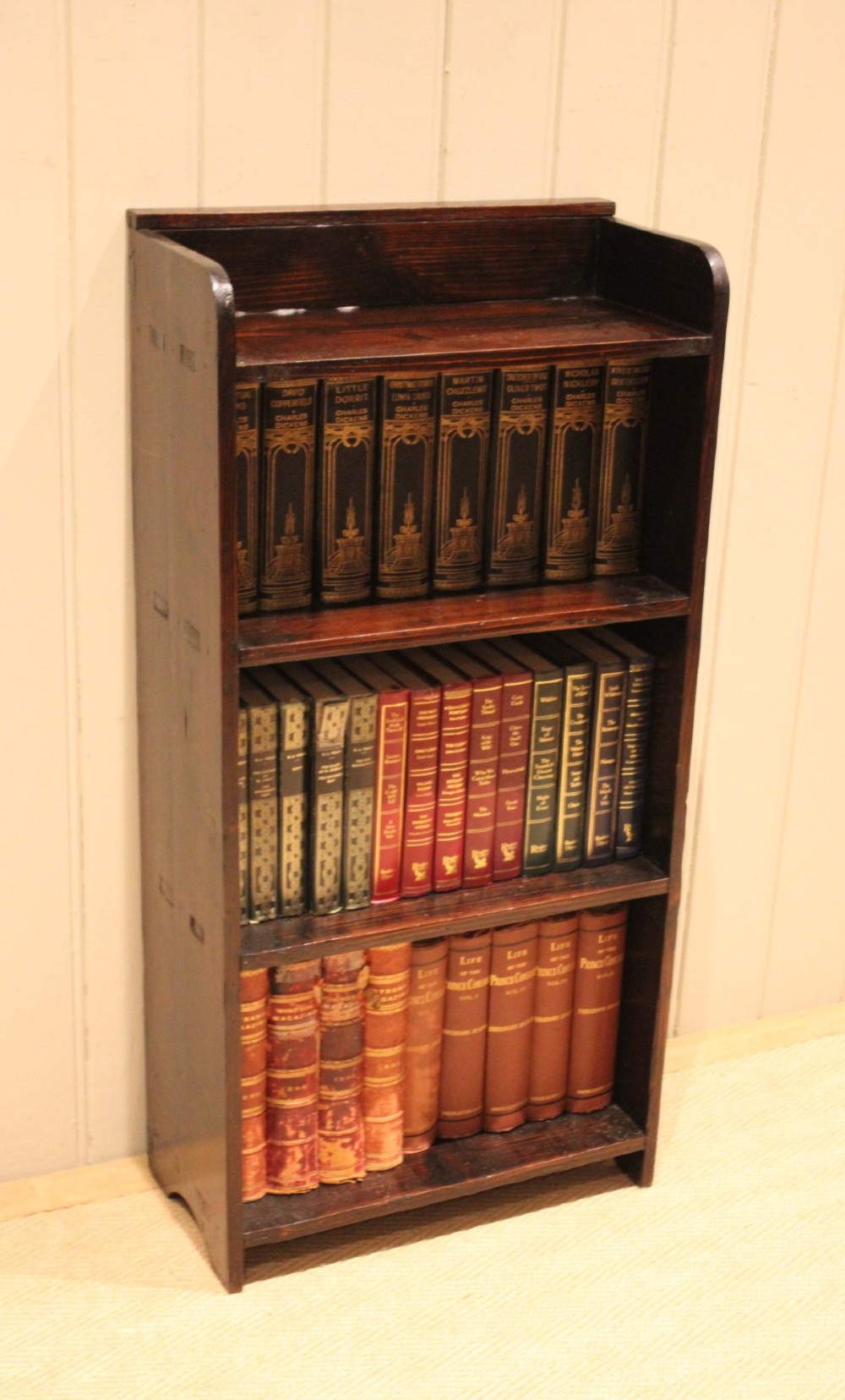A small dark pine open bookcase featuring three fixed shelves, solid side panels with visible tenon joinery, and a simple shaped gallery top. View VII