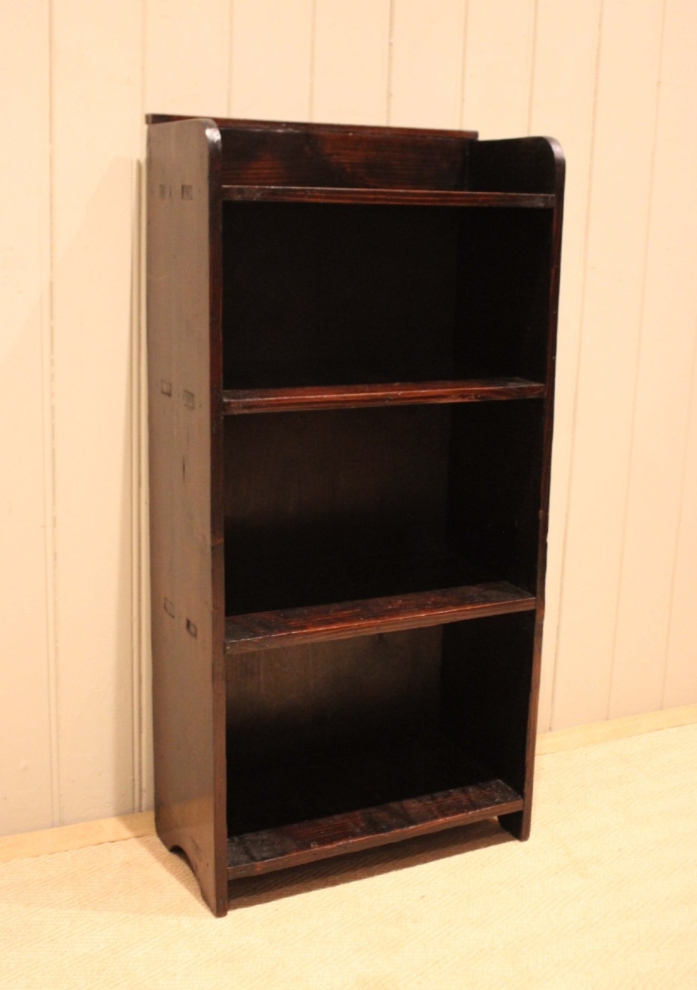 A small dark pine open bookcase featuring three fixed shelves, solid side panels with visible tenon joinery, and a simple shaped gallery top. View VIII