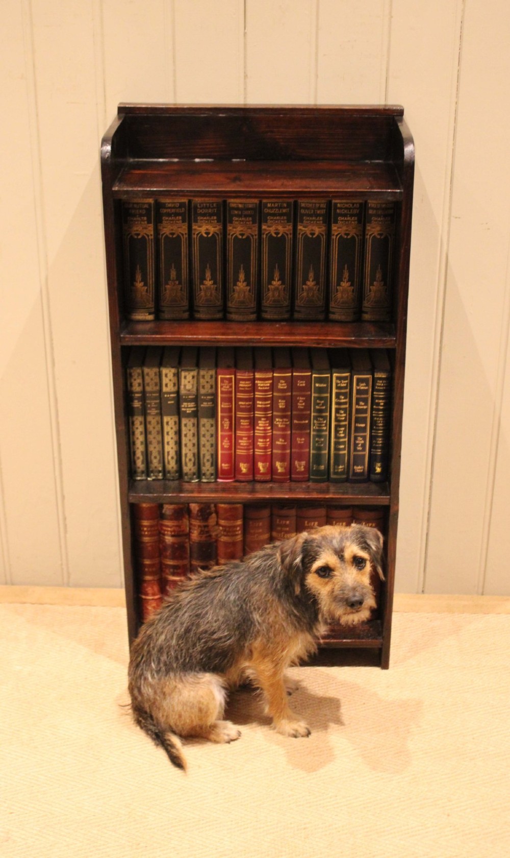 A small dark pine open bookcase featuring three fixed shelves, solid side panels with visible tenon joinery, and a simple shaped gallery top. View IX