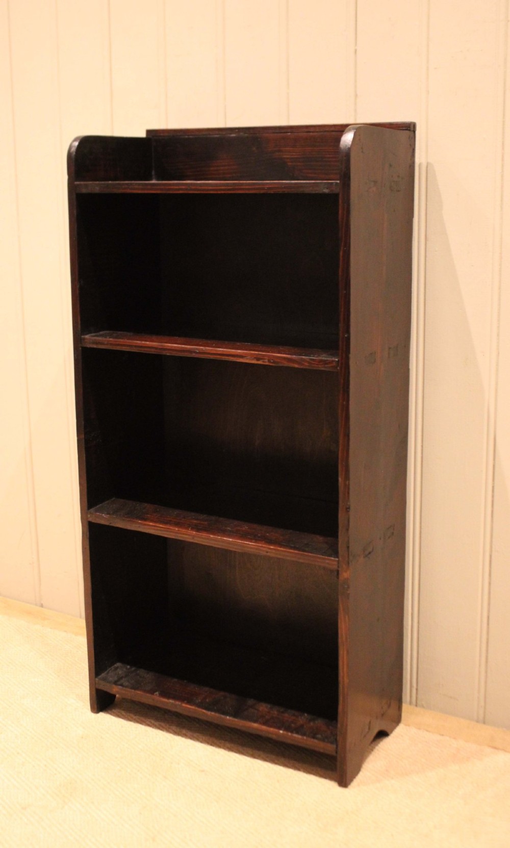A small dark pine open bookcase featuring three fixed shelves, solid side panels with visible tenon joinery, and a simple shaped gallery top. View X
