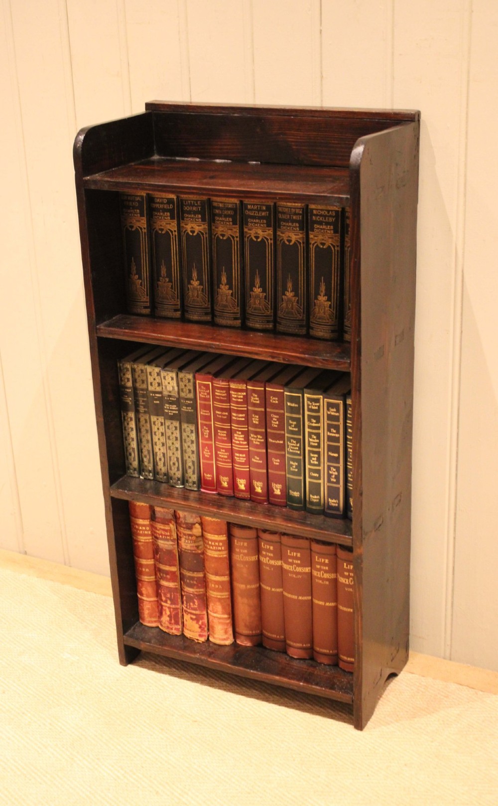 A small dark pine open bookcase featuring three fixed shelves, solid side panels with visible tenon joinery, and a simple shaped gallery top. View XI