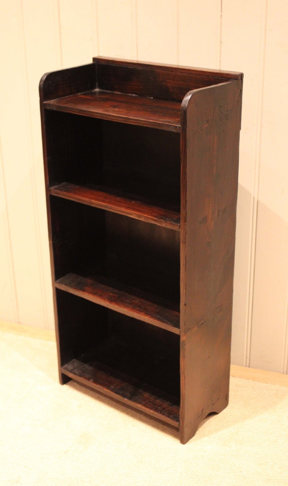 A small dark pine open bookcase featuring three fixed shelves, solid side panels with visible tenon joinery, and a simple shaped gallery top. View XII