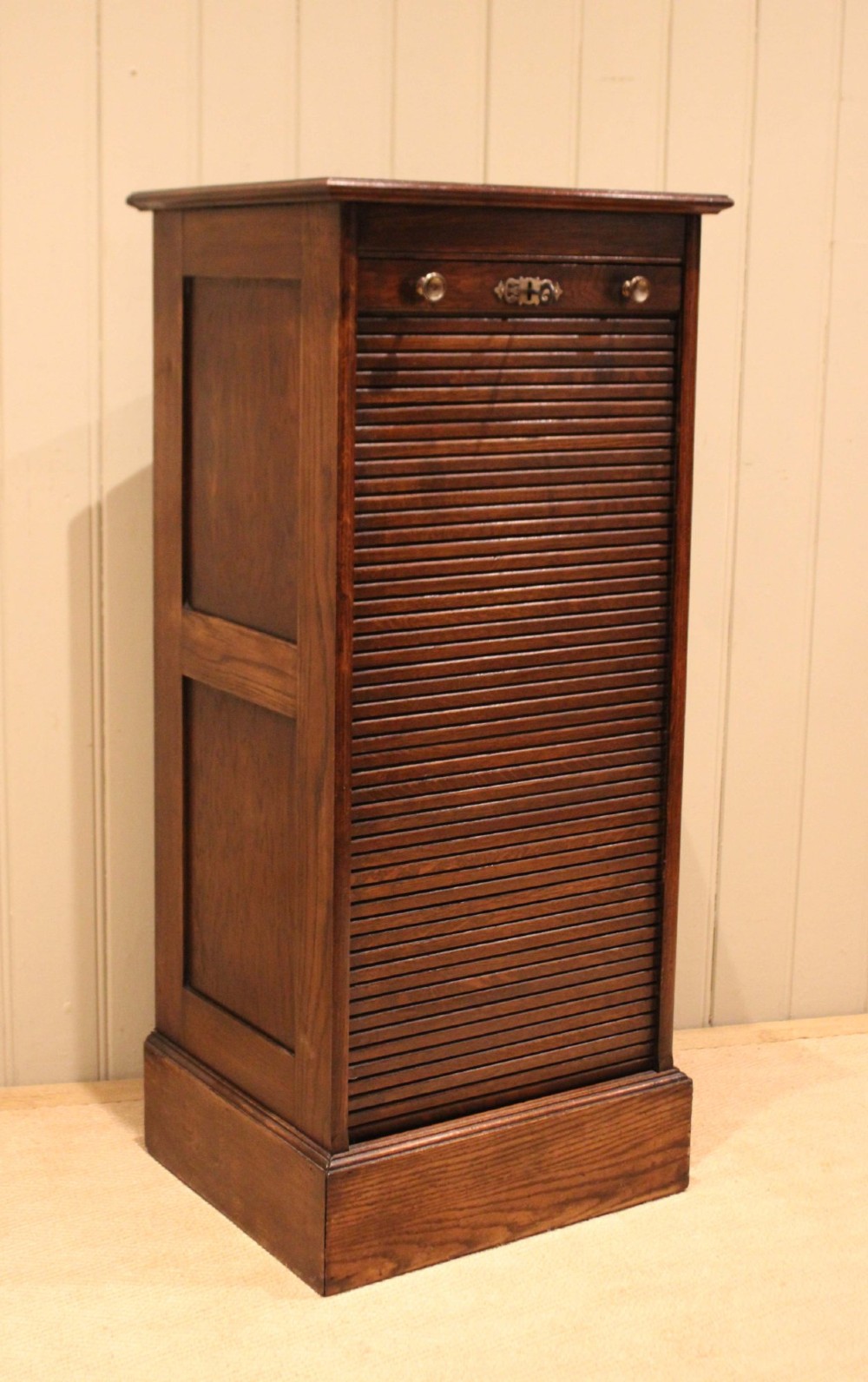 A tall dark oak single-column filing cabinet featuring a slatted wooden tambour front with top brass pull knobs and a keyhole escutcheon.