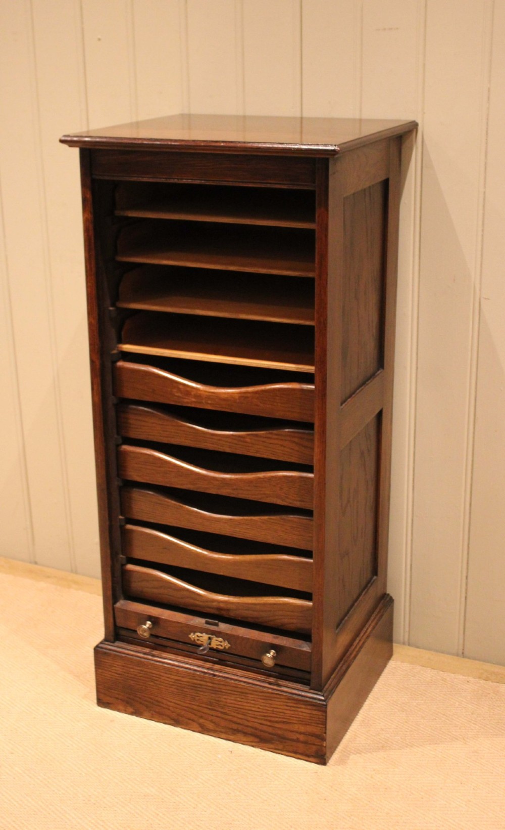 A tall dark oak single-column filing cabinet featuring a slatted wooden tambour front with top brass pull knobs and a keyhole escutcheon. View II