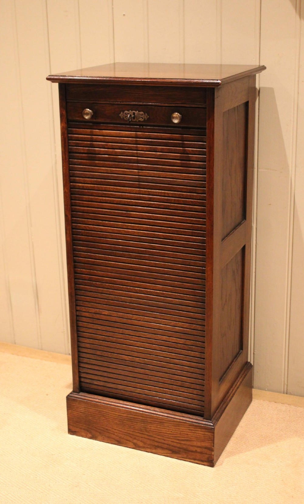 A tall dark oak single-column filing cabinet featuring a slatted wooden tambour front with top brass pull knobs and a keyhole escutcheon. View III
