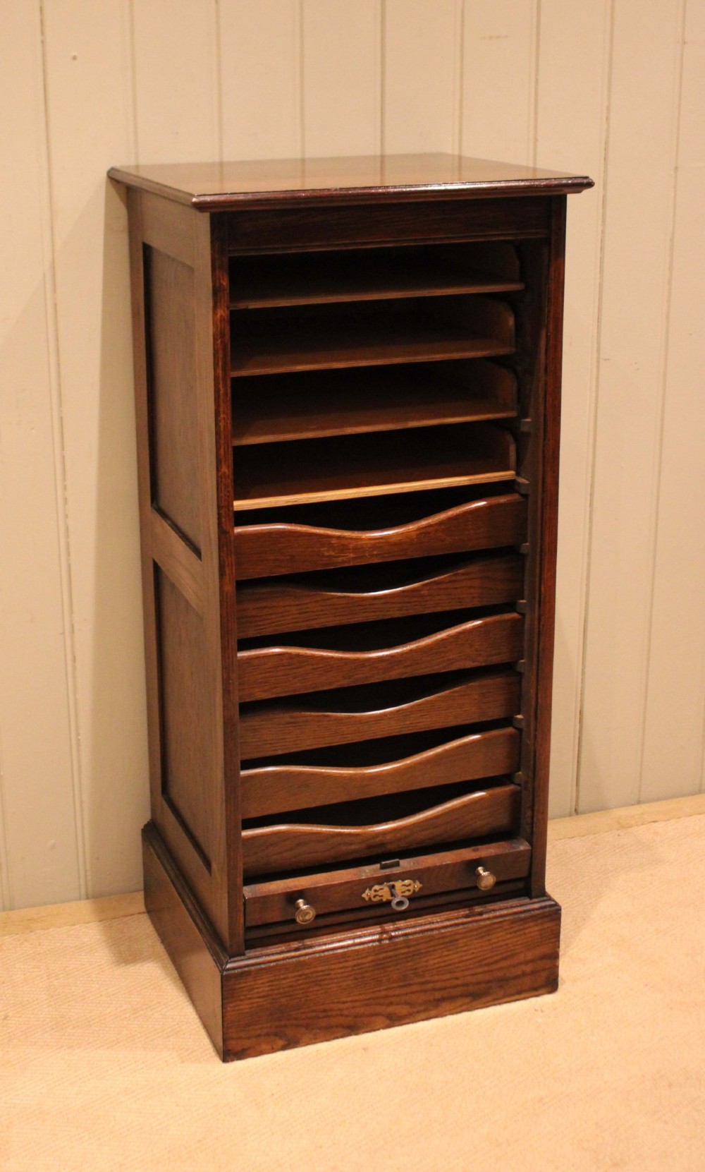 A tall dark oak single-column filing cabinet featuring a slatted wooden tambour front with top brass pull knobs and a keyhole escutcheon. View IV