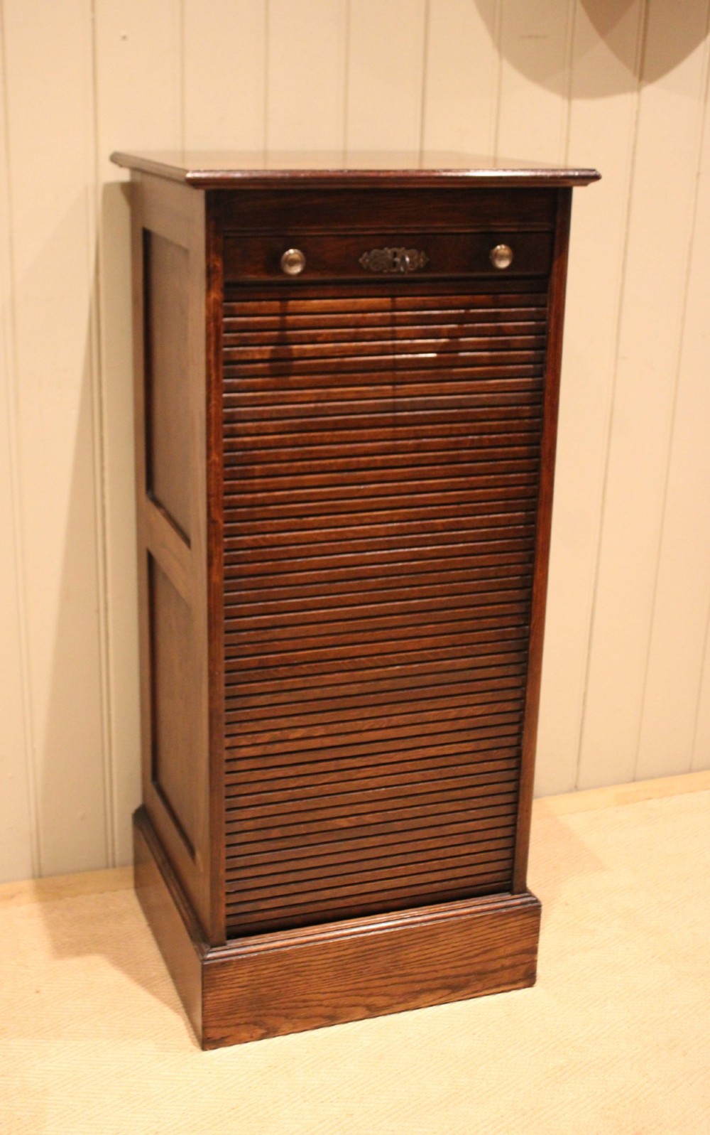 A tall dark oak single-column filing cabinet featuring a slatted wooden tambour front with top brass pull knobs and a keyhole escutcheon. View VII