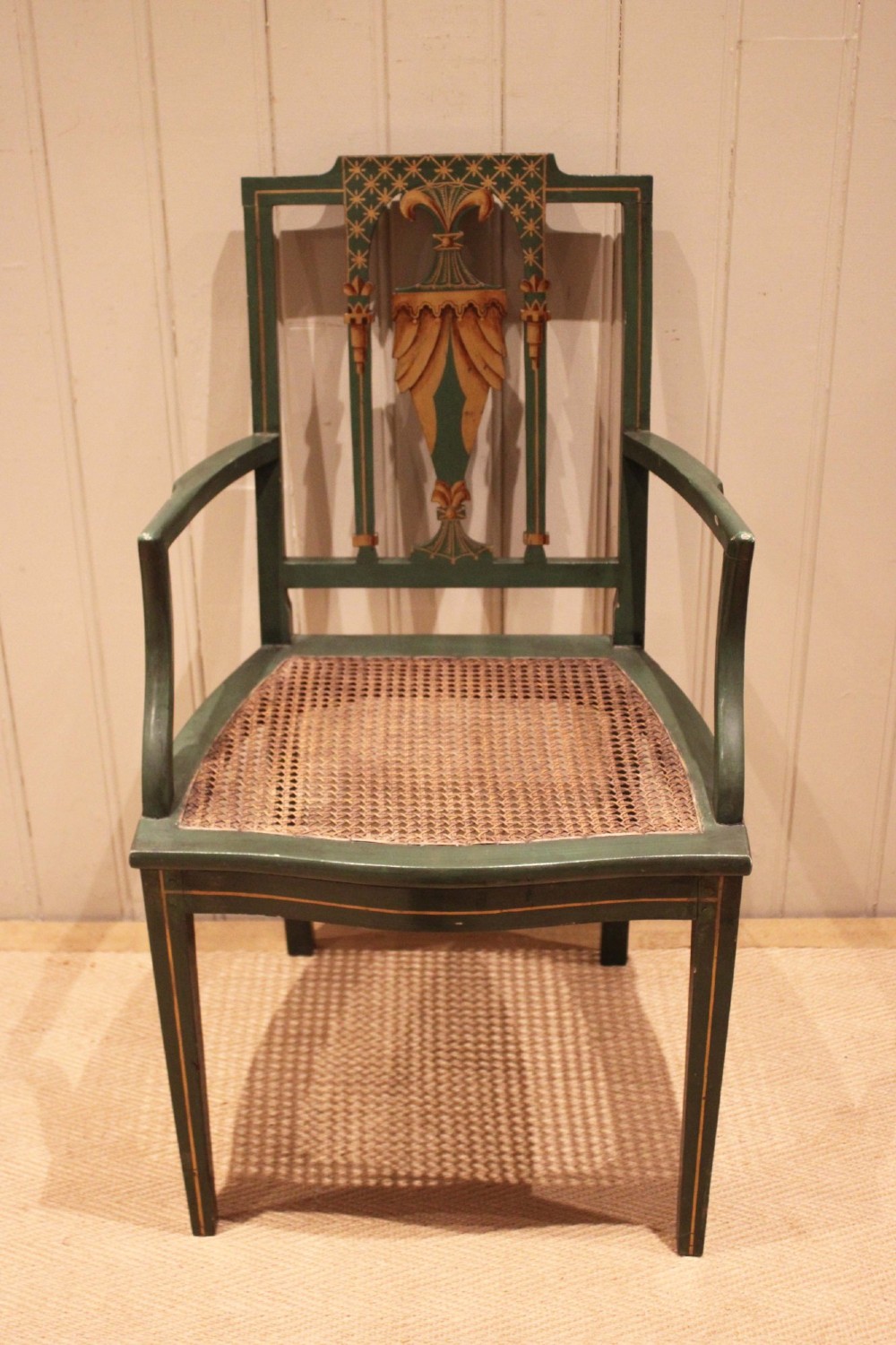 A dark green painted Hepplewhite style armchair featuring a woven cane seat, curved armrests, and hand-painted neoclassical motifs on the backrest.