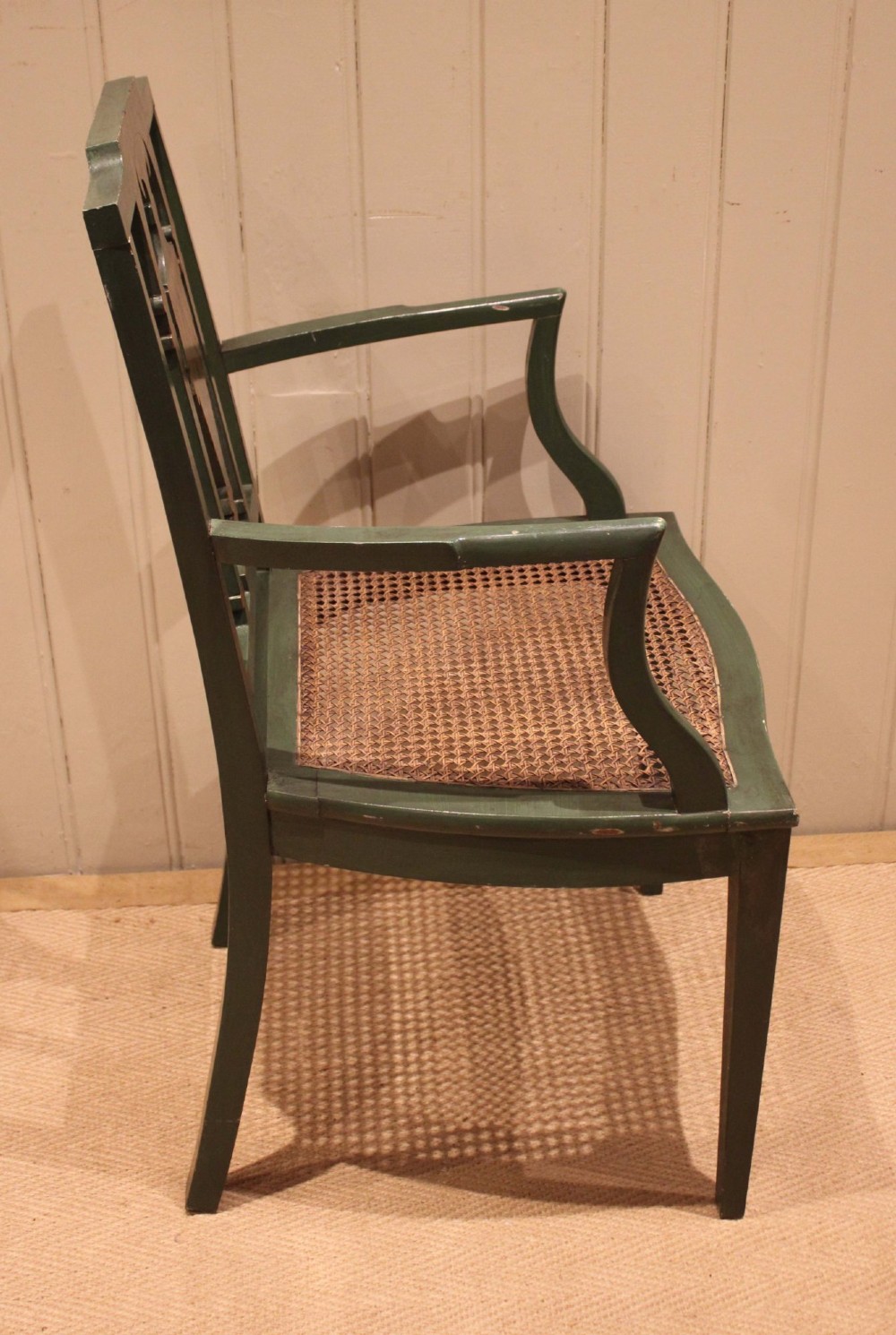 A dark green painted Hepplewhite style armchair featuring a woven cane seat, curved armrests, and hand-painted neoclassical motifs on the backrest. View X