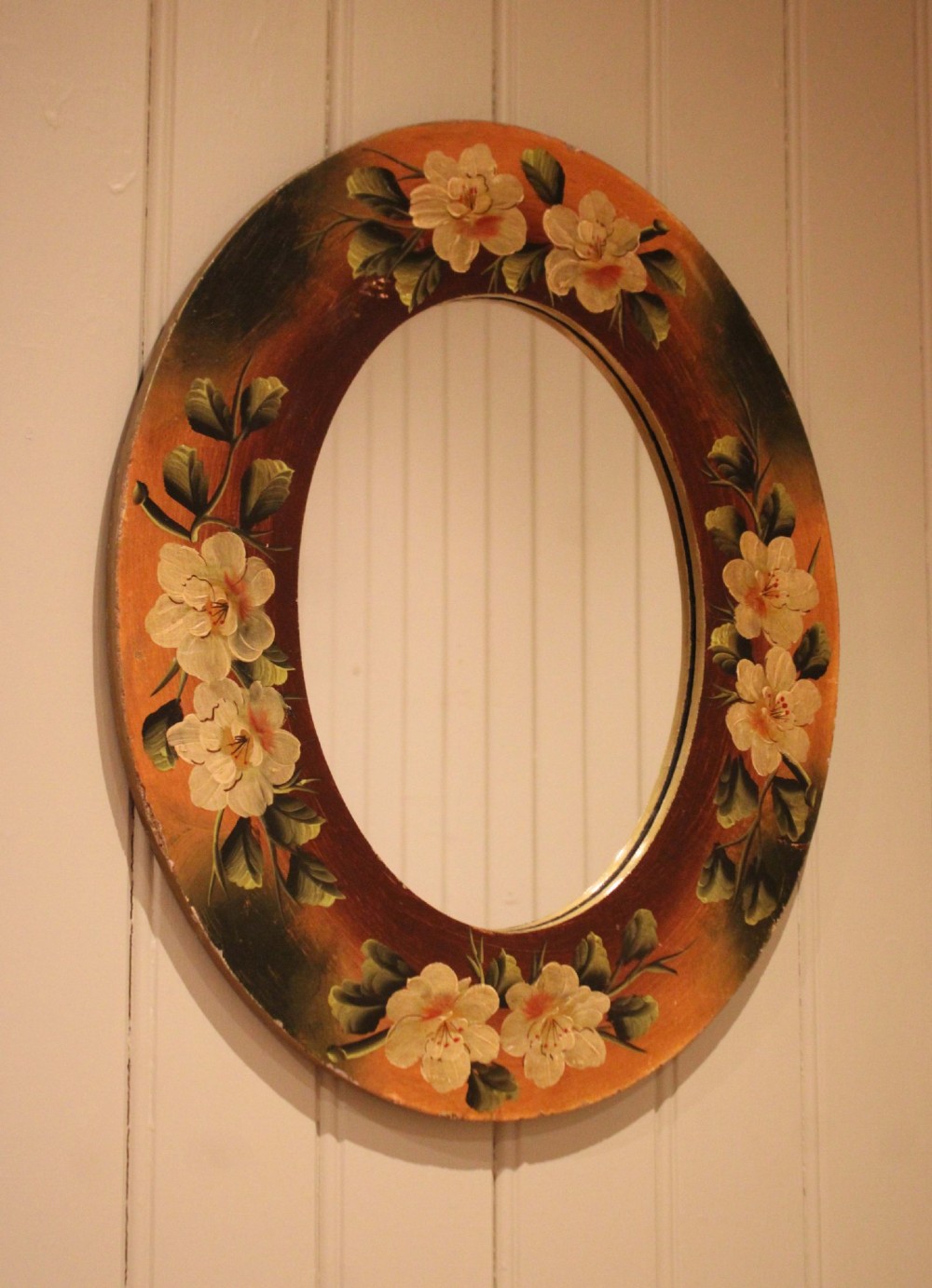 An oval wooden wall mirror featuring hand-painted cream floral blossoms and green foliage on an amber-toned frame. View II
