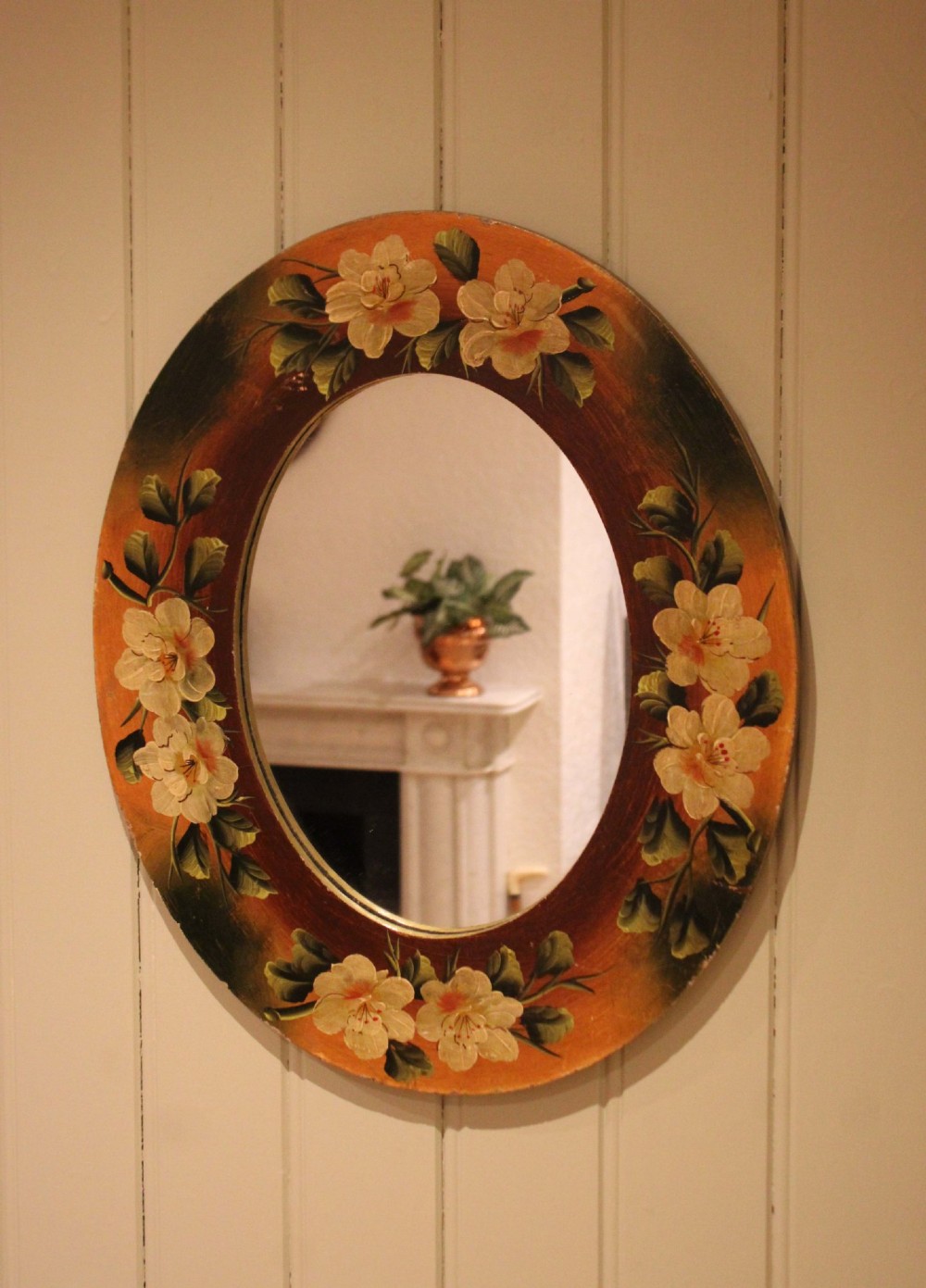 An oval wooden wall mirror featuring hand-painted cream floral blossoms and green foliage on an amber-toned frame. View III