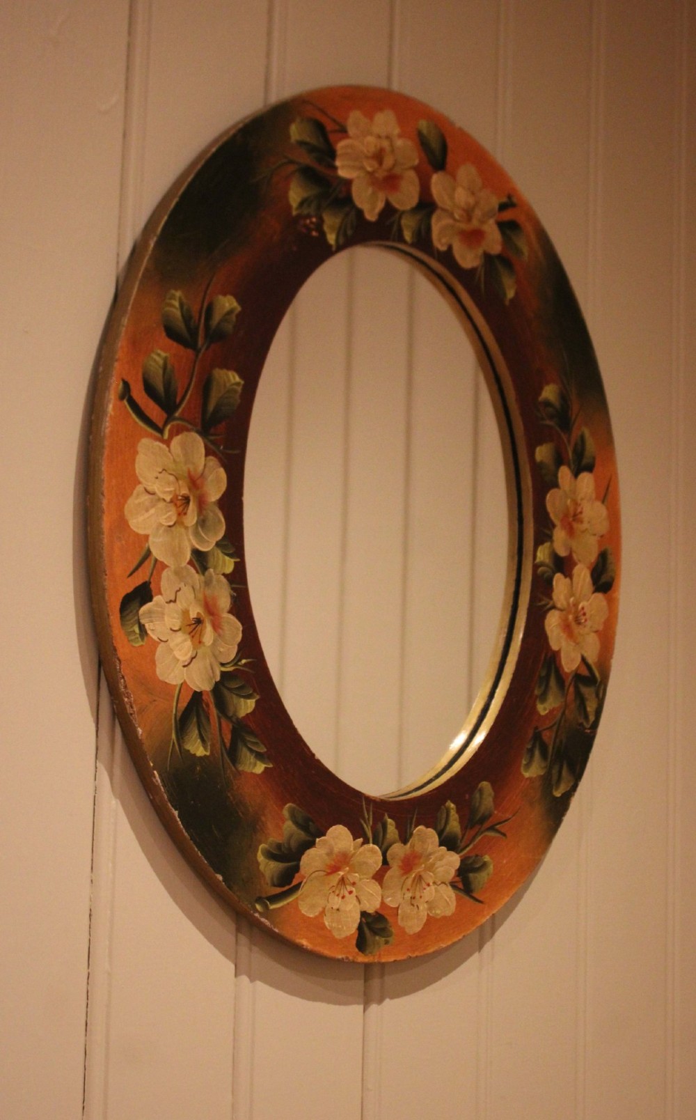 An oval wooden wall mirror featuring hand-painted cream floral blossoms and green foliage on an amber-toned frame. View IV