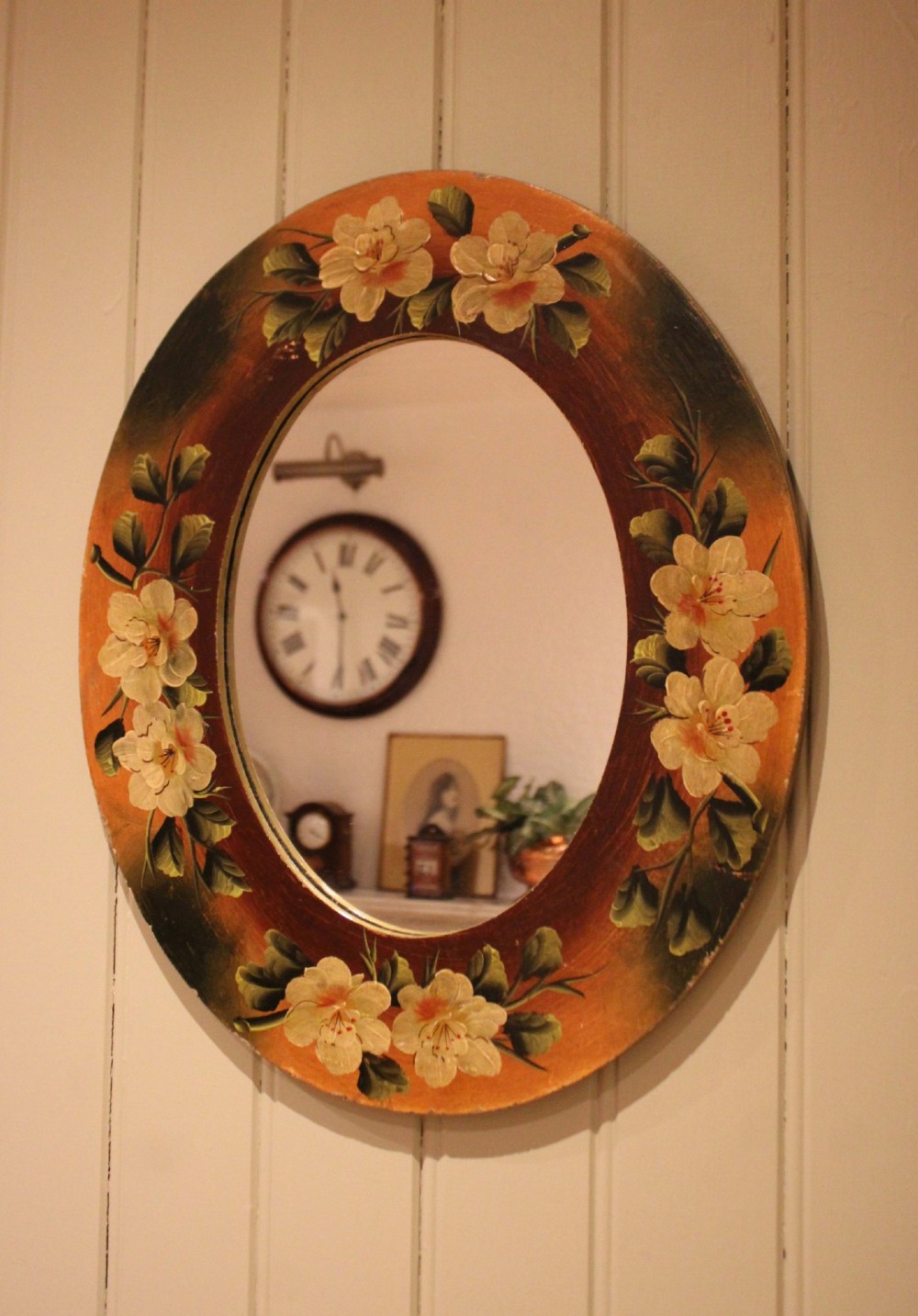 An oval wooden wall mirror featuring hand-painted cream floral blossoms and green foliage on an amber-toned frame. View V