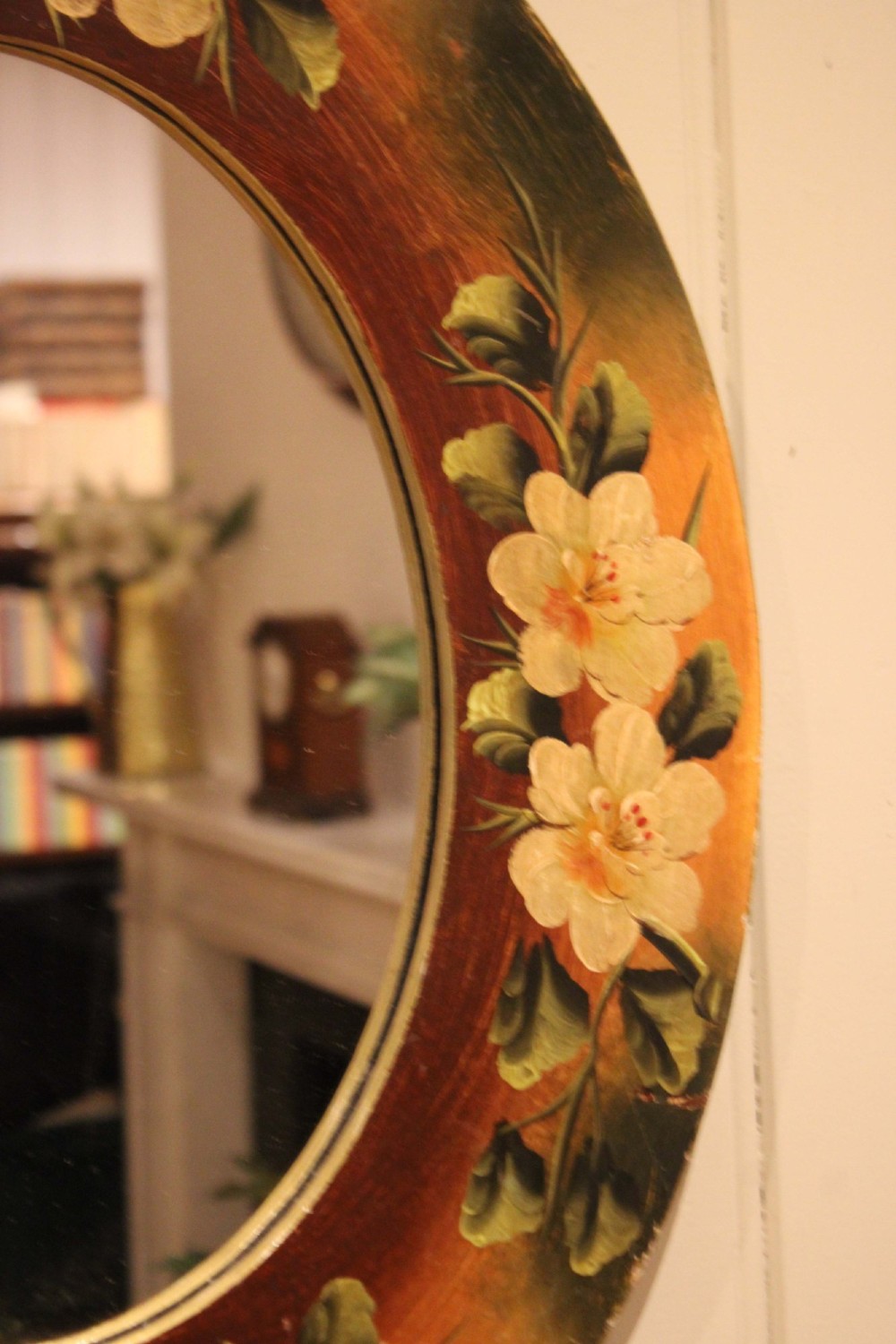 An oval wooden wall mirror featuring hand-painted cream floral blossoms and green foliage on an amber-toned frame. View VI