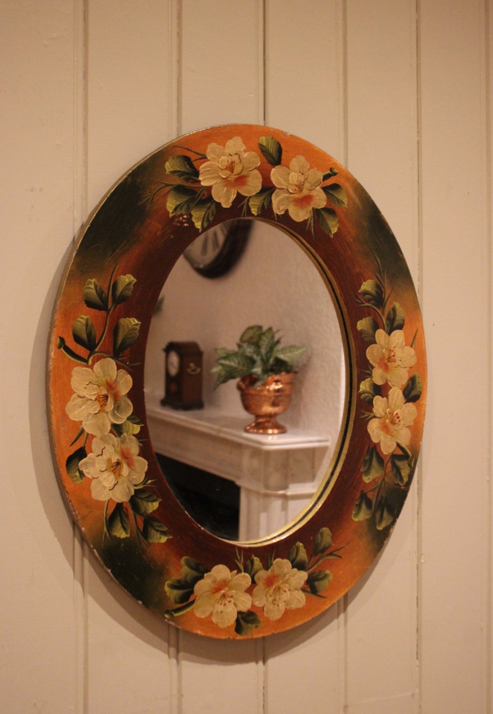 An oval wooden wall mirror featuring hand-painted cream floral blossoms and green foliage on an amber-toned frame. View VII