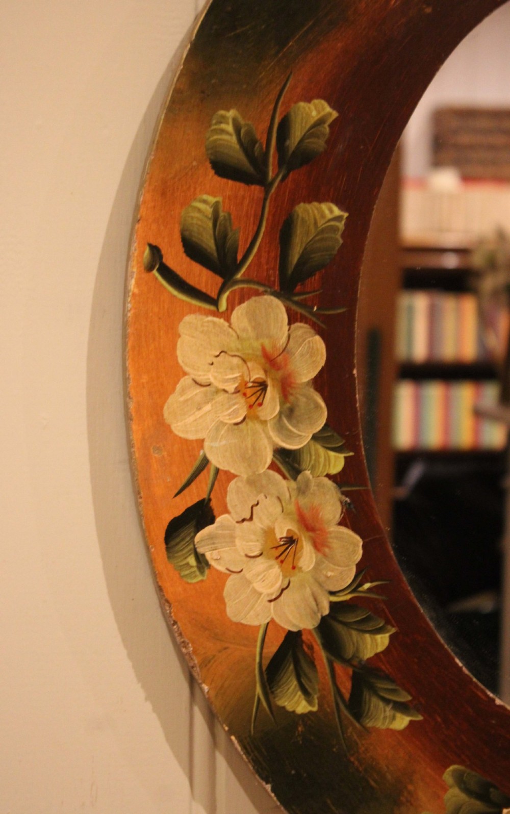 An oval wooden wall mirror featuring hand-painted cream floral blossoms and green foliage on an amber-toned frame. View VIII
