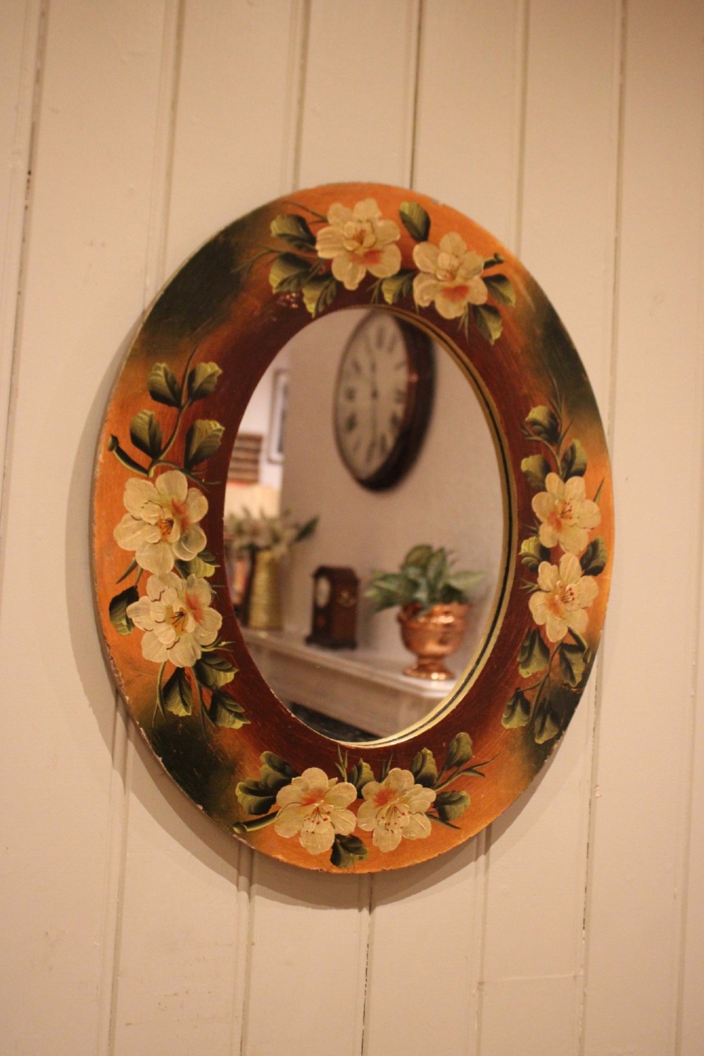 An oval wooden wall mirror featuring hand-painted cream floral blossoms and green foliage on an amber-toned frame. View IX