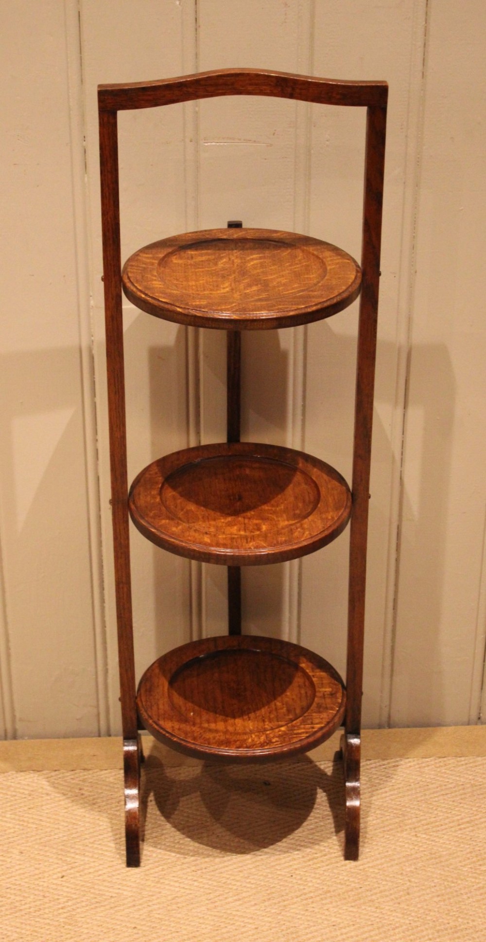 An Edwardian oak three-tier folding cake stand featuring circular recessed shelves and an arched carrying handle.