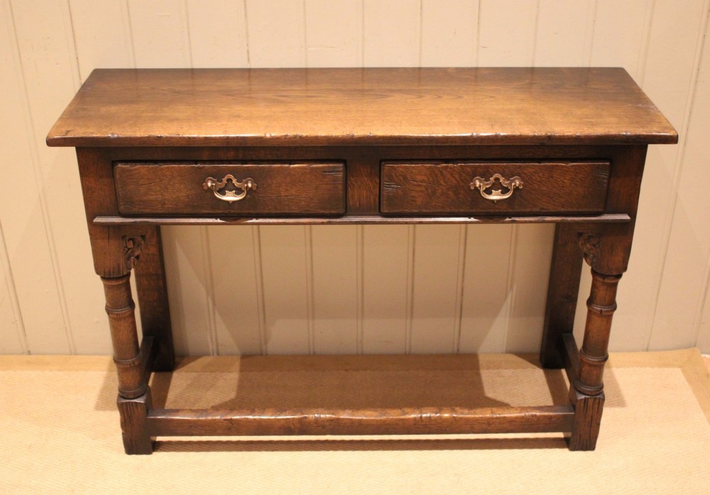 A solid oak server table featuring a rectangular top, two drawers with brass swan-neck handles, turned front legs, and a lower stretcher base.